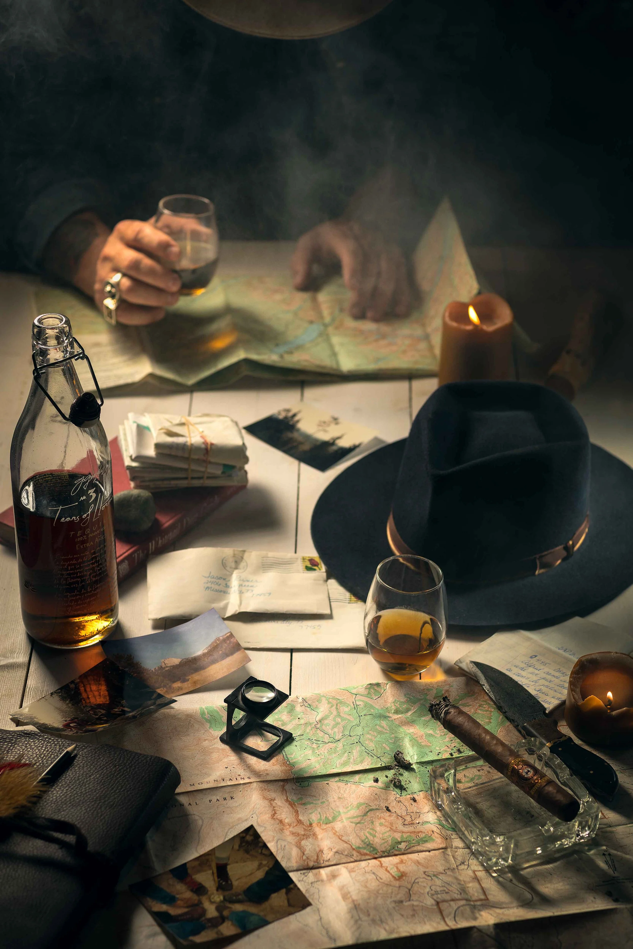 A table with maps, photographs, a cigar, a hat, a flashlight, a whiskey glass, and a bottle of whiskey, suggesting outdoor exploration or planning.