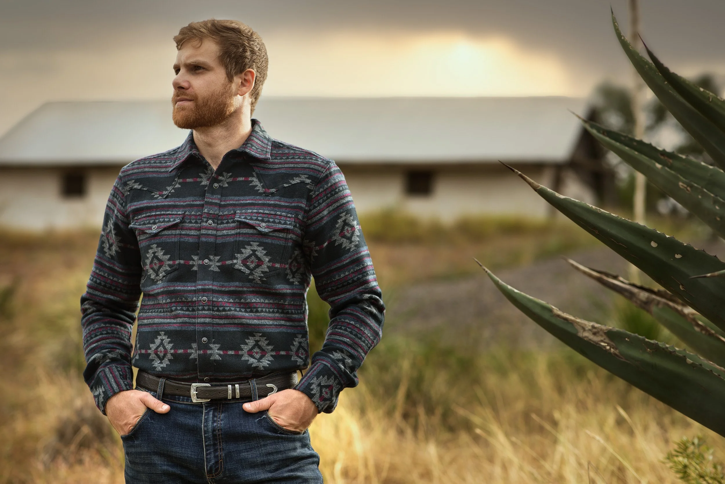 A man with a beard and short brown hair standing outdoors with his hands in his pockets, wearing a patterned flannel shirt and blue jeans, near a large aloe vera plant and a building in the background during late afternoon or early evening.