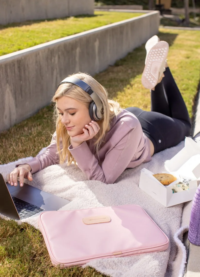 Young woman with headphones lying on her stomach on a blanket, outdoors, using a laptop, with a box of food nearby.