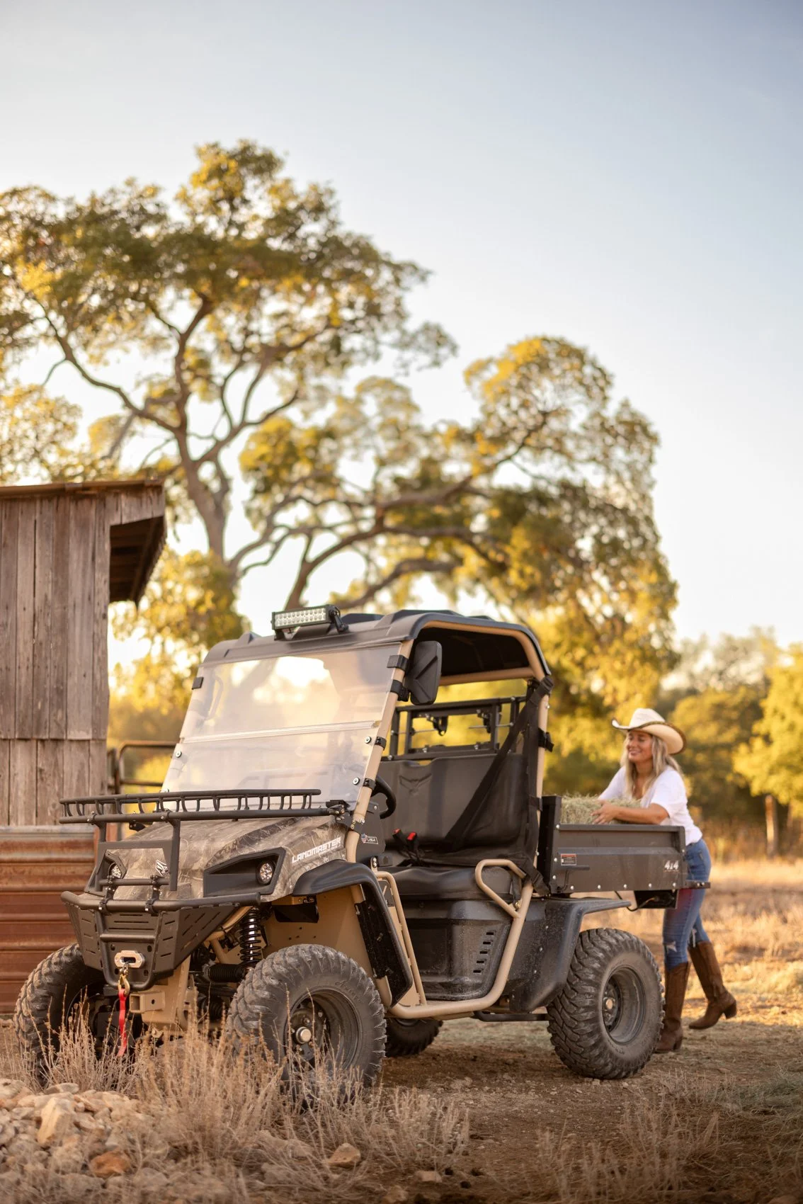 A woman in cowboy boots and a cowboy hat standing next to an off-road utility vehicle in a rural outdoor setting with trees and a wooden shed, during sunset.
