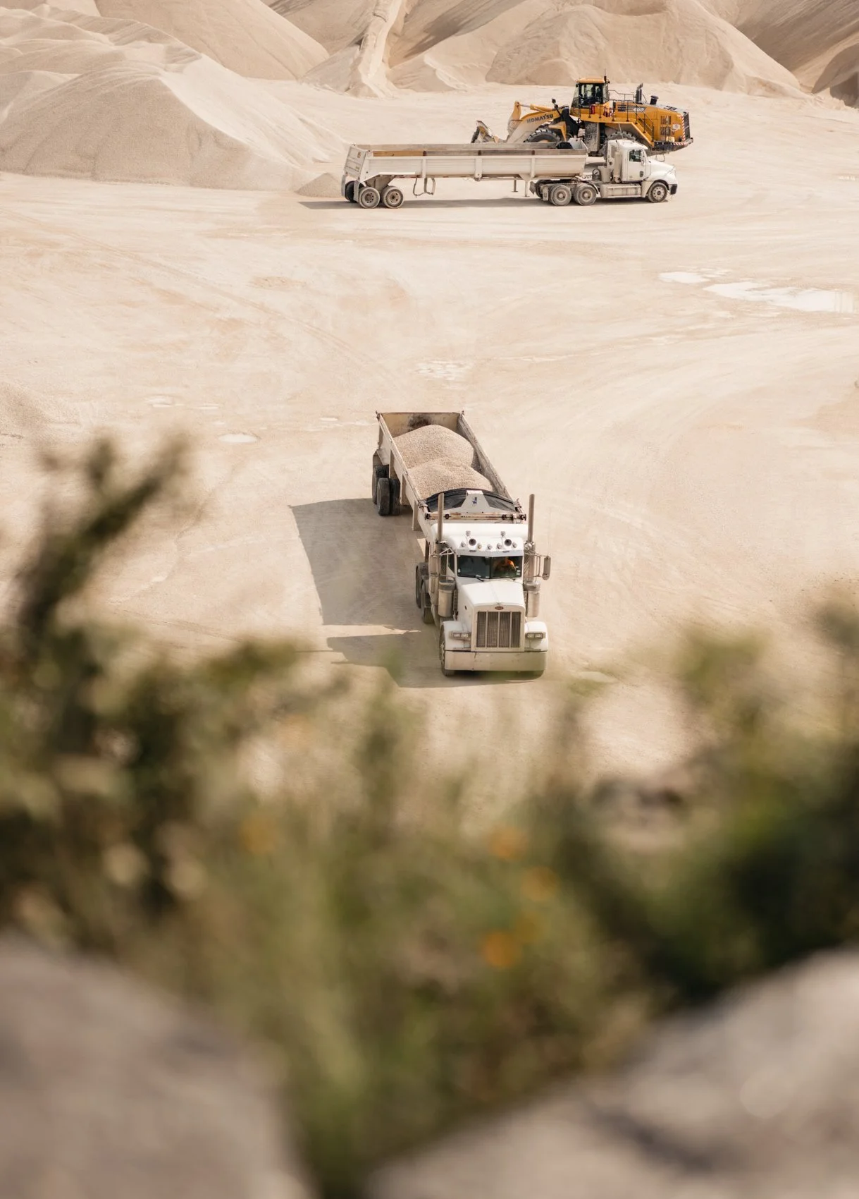 Dump trucks transporting sand or gravel in a quarry or construction site with hills and machinery in the background.