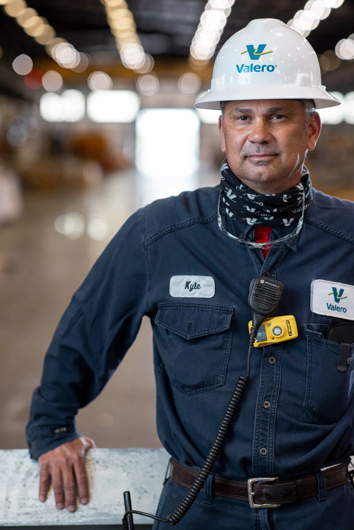 A man wearing a white hard hat with the Valero logo, a navy blue work uniform with a name tag that says 'Kyle', a radio, and safety gear, standing in an industrial or warehouse setting.