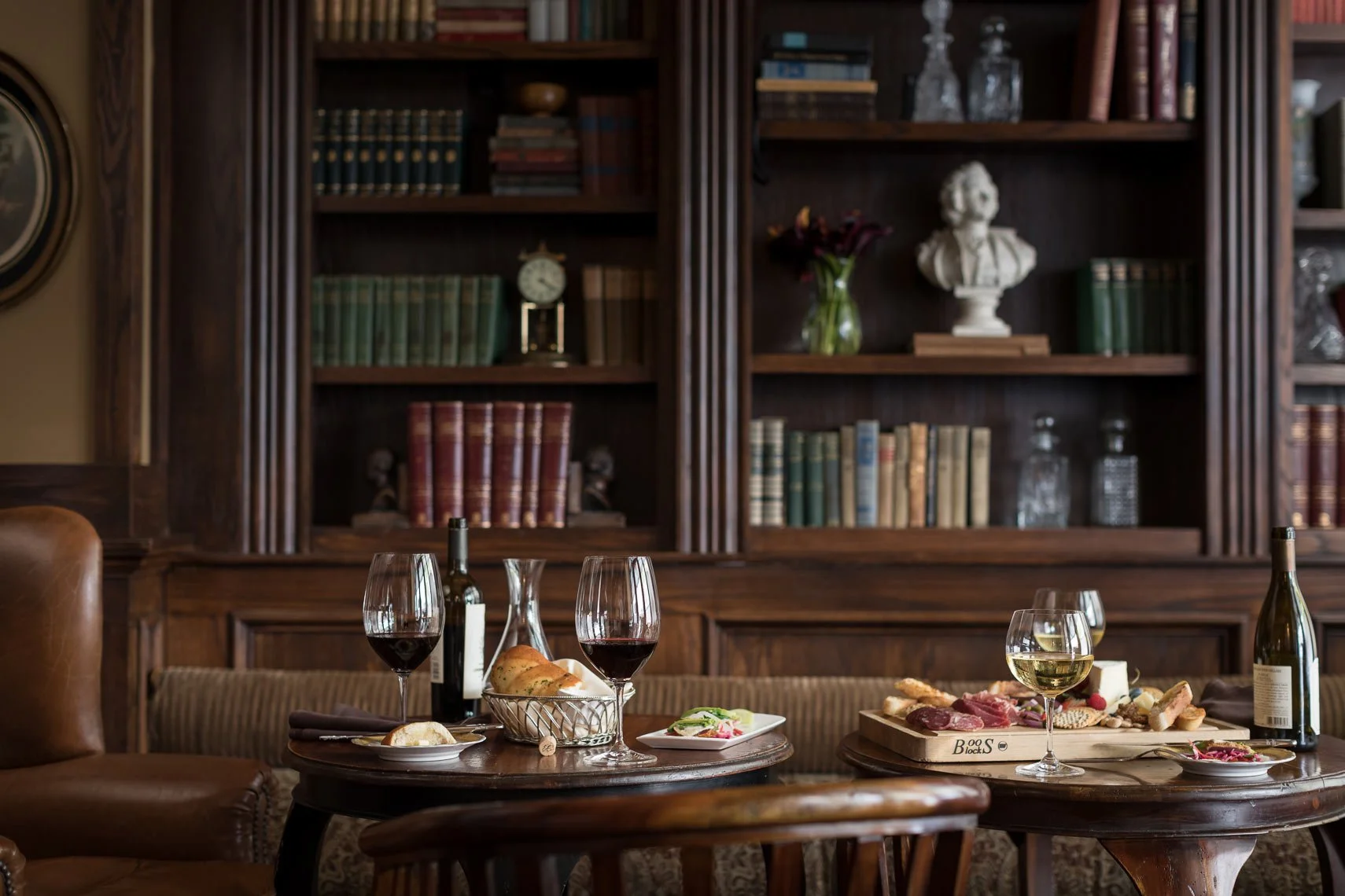 A cozy dining room with a wooden table set with wine glasses, a wine bottle, plates of food, and a cheese platter, with a large wooden bookshelf filled with books and decorative items in the background.