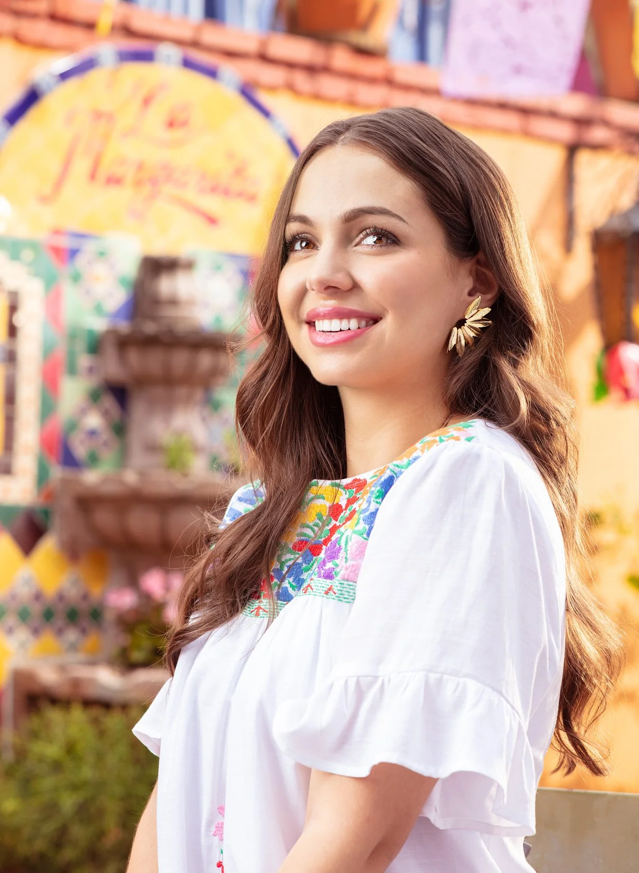 A young woman with light skin, long brown hair, and wearing a white blouse with colorful embroidery. She is smiling and looking slightly upward. The background features vibrant colorful decorations and blurred elements that suggest an outdoor festive