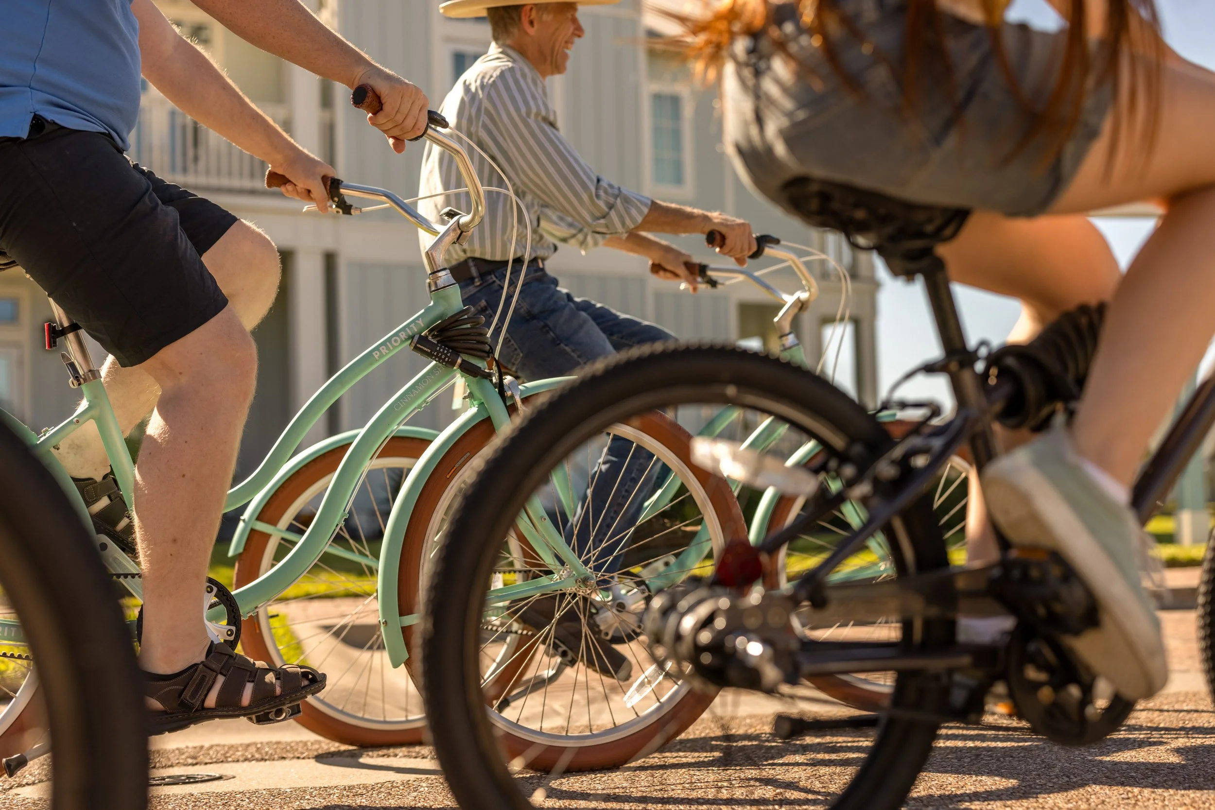 Close-up of a group of people riding bicycles on a sidewalk in front of a residential building, with an elderly woman smiling in the background.