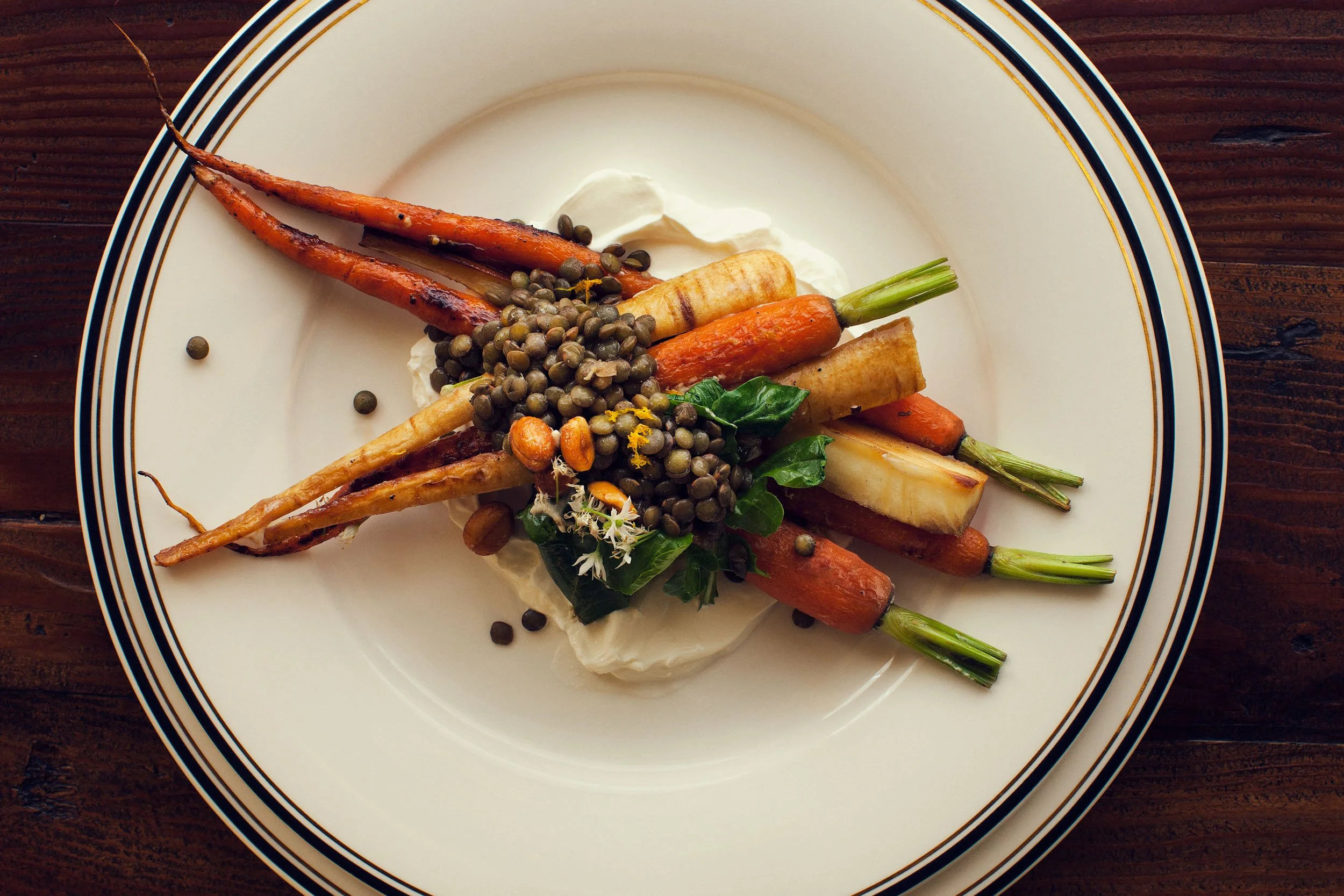 A plate of roasted carrots, parsnips, and green beans, topped with green peppercorns and garnished with herbs, on a white plate with a navy and gold rim, wooden table background.