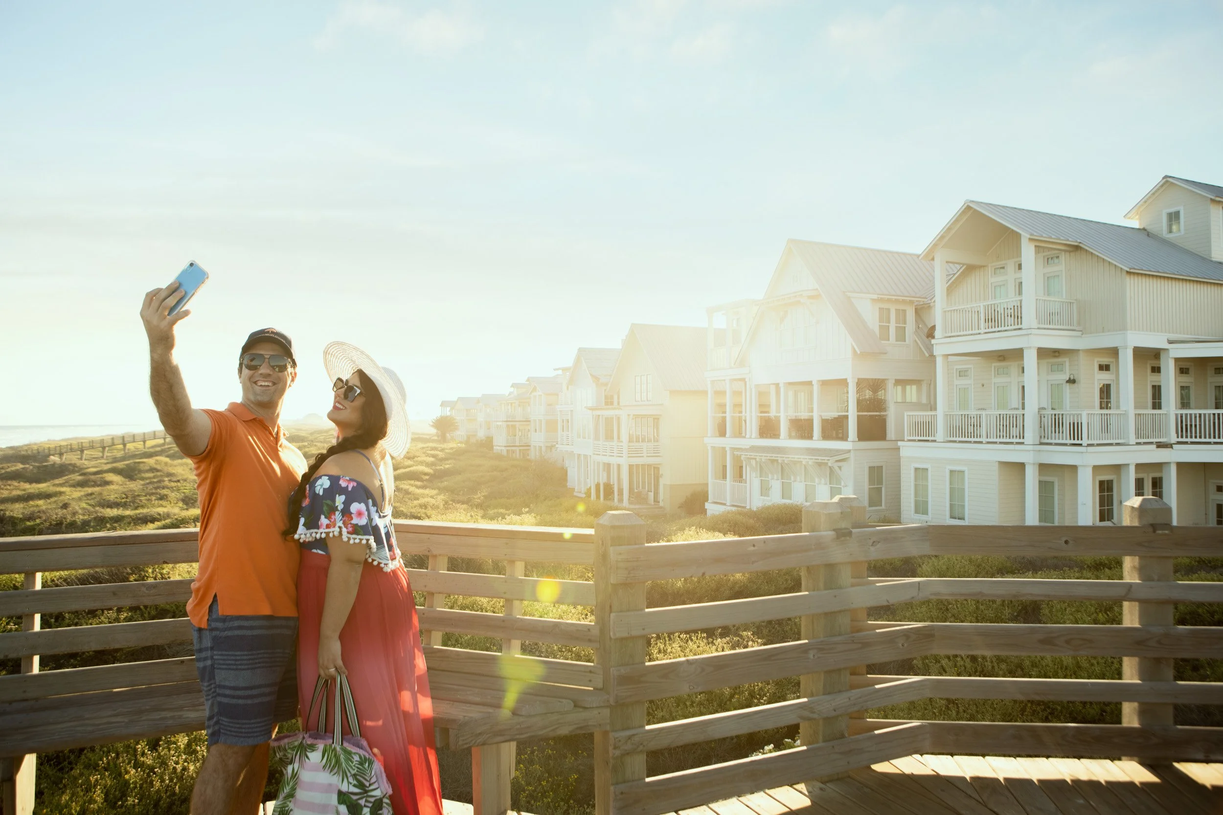 A man and woman taking a selfie outdoors near beach houses at sunset, with the man holding a phone and both smiling.