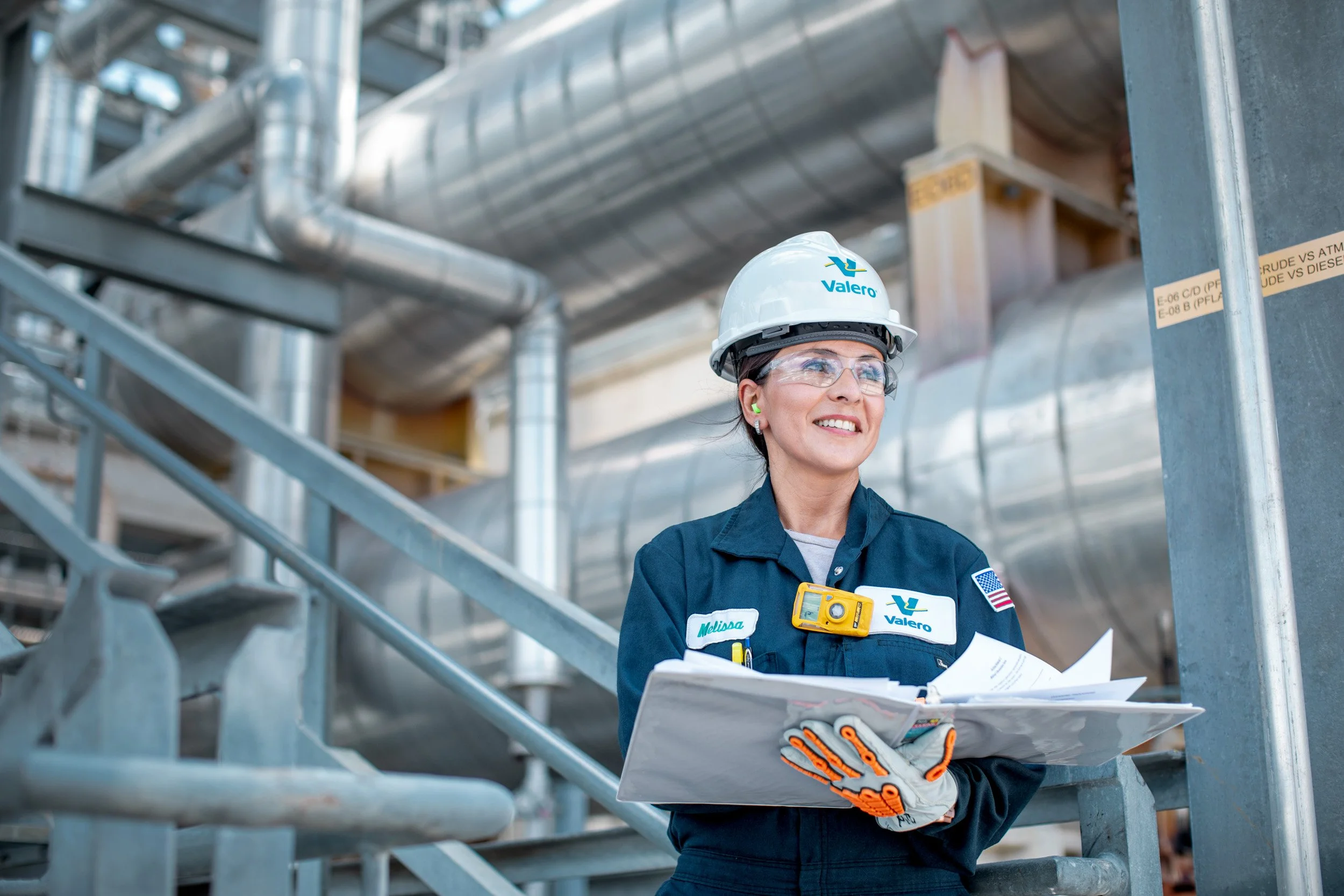 A woman in a blue work uniform, safety glasses, and a hard hat labeled Valero, stands on industrial stairs holding papers and a small yellow device, smiling in an industrial setting with pipes and metal structures.