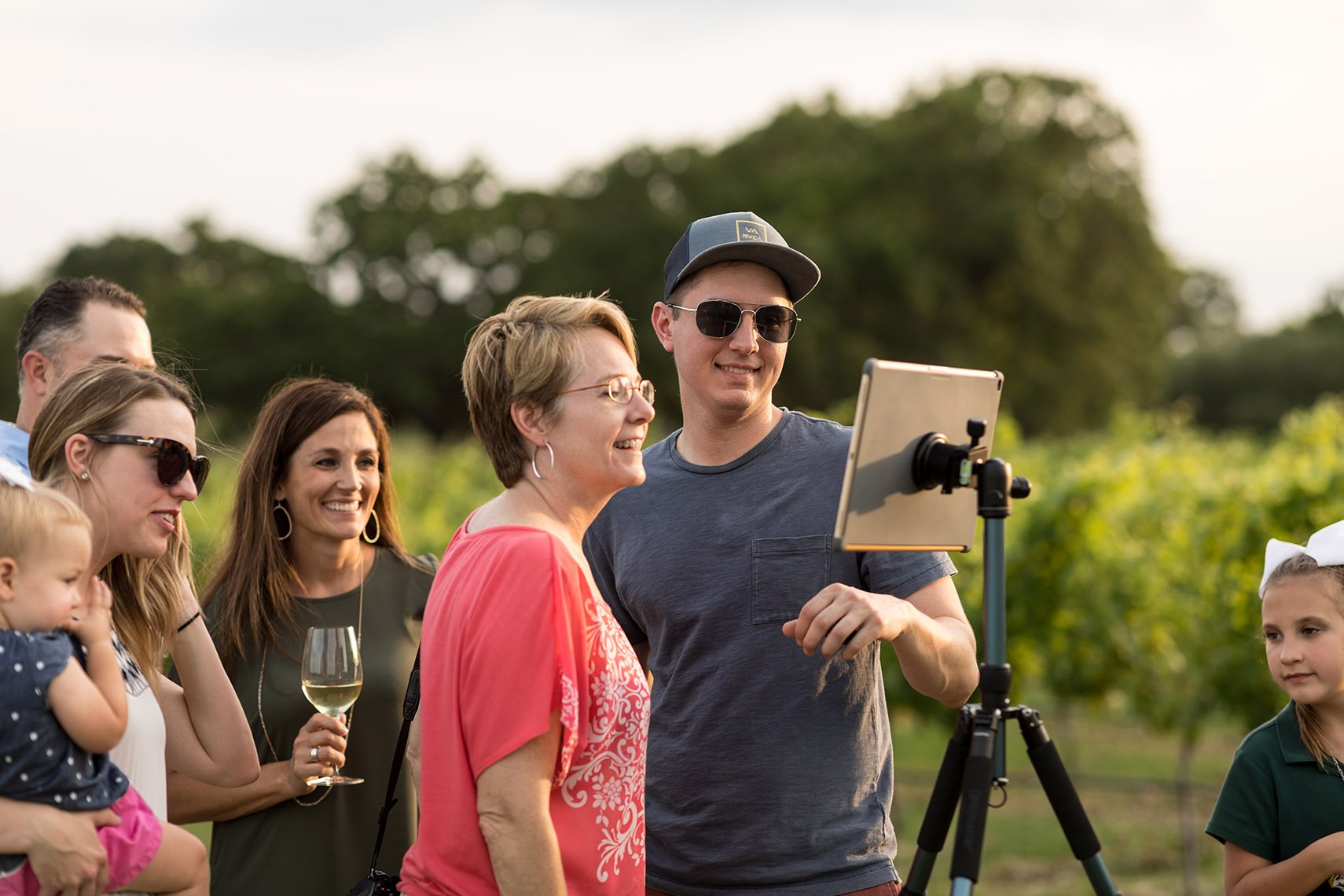 A group of people, including children and adults, are gathered outdoors during the daytime, looking at a tablet on a tripod. They are smiling and seem to be enjoying a social event or celebration.