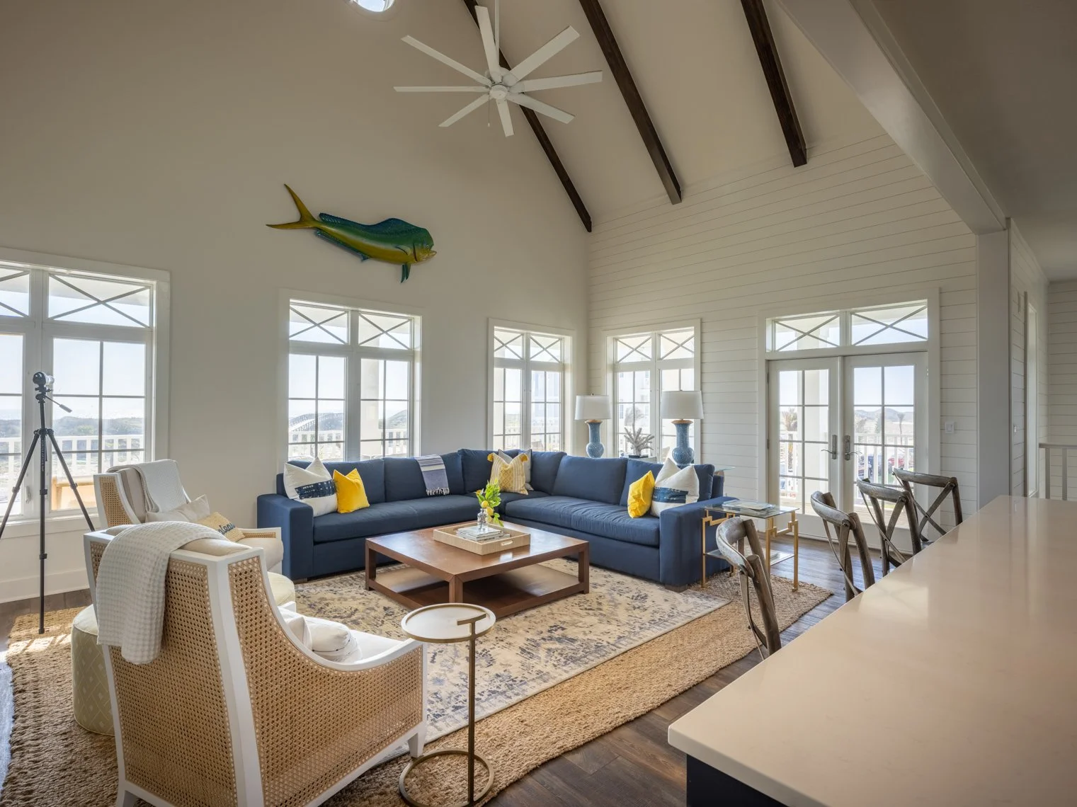 A spacious living room with large windows, a navy sectional sofa with yellow and white pillows, a wooden coffee table, and a beige armchair. There are two table lamps on side tables, a patterned area rug, and a decorative fish wall sculpture. The roo