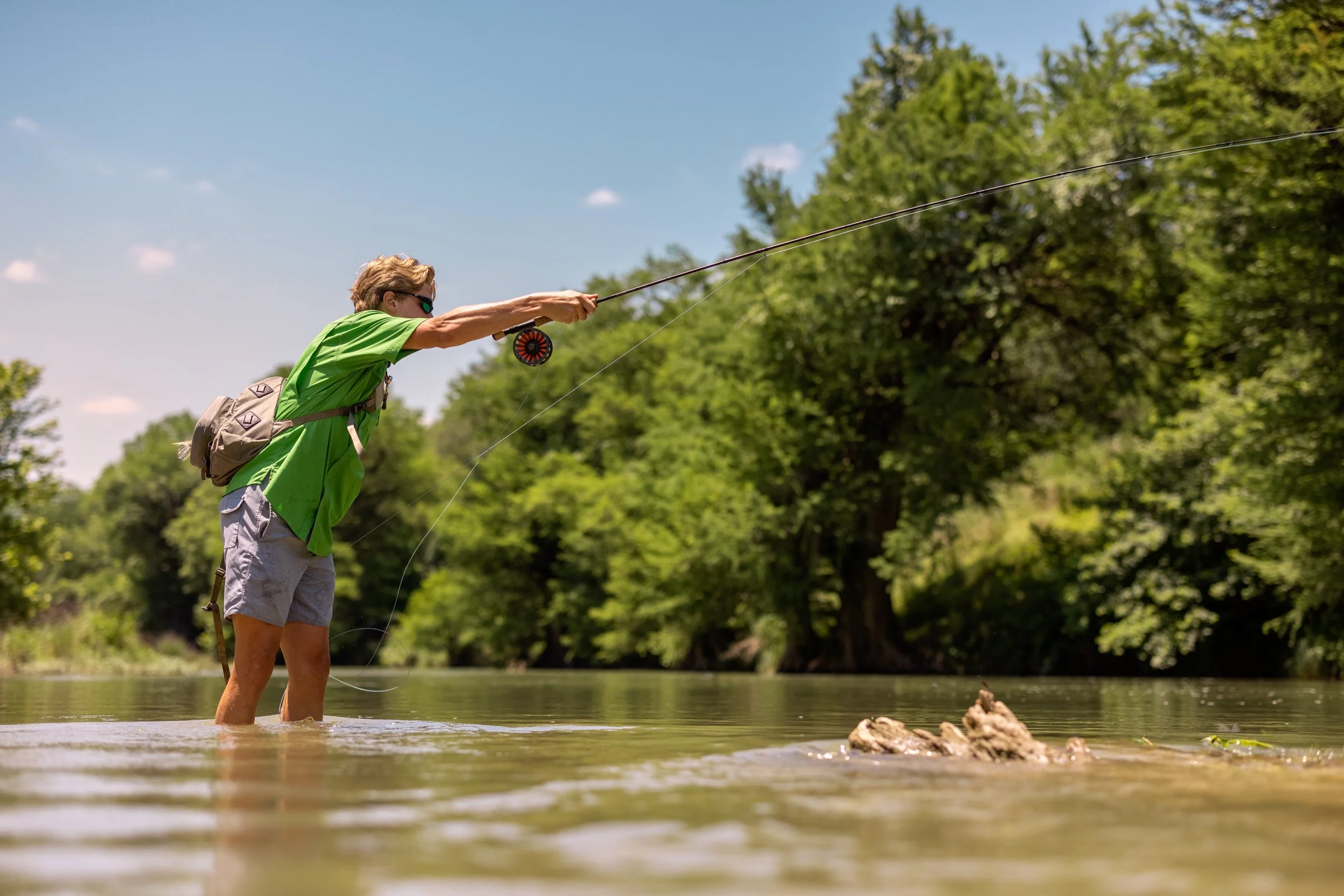 A person standing in a shallow river, fishing with a rod, wearing sunglasses, a green shirt, and gray shorts, surrounded by green trees under a bright blue sky.