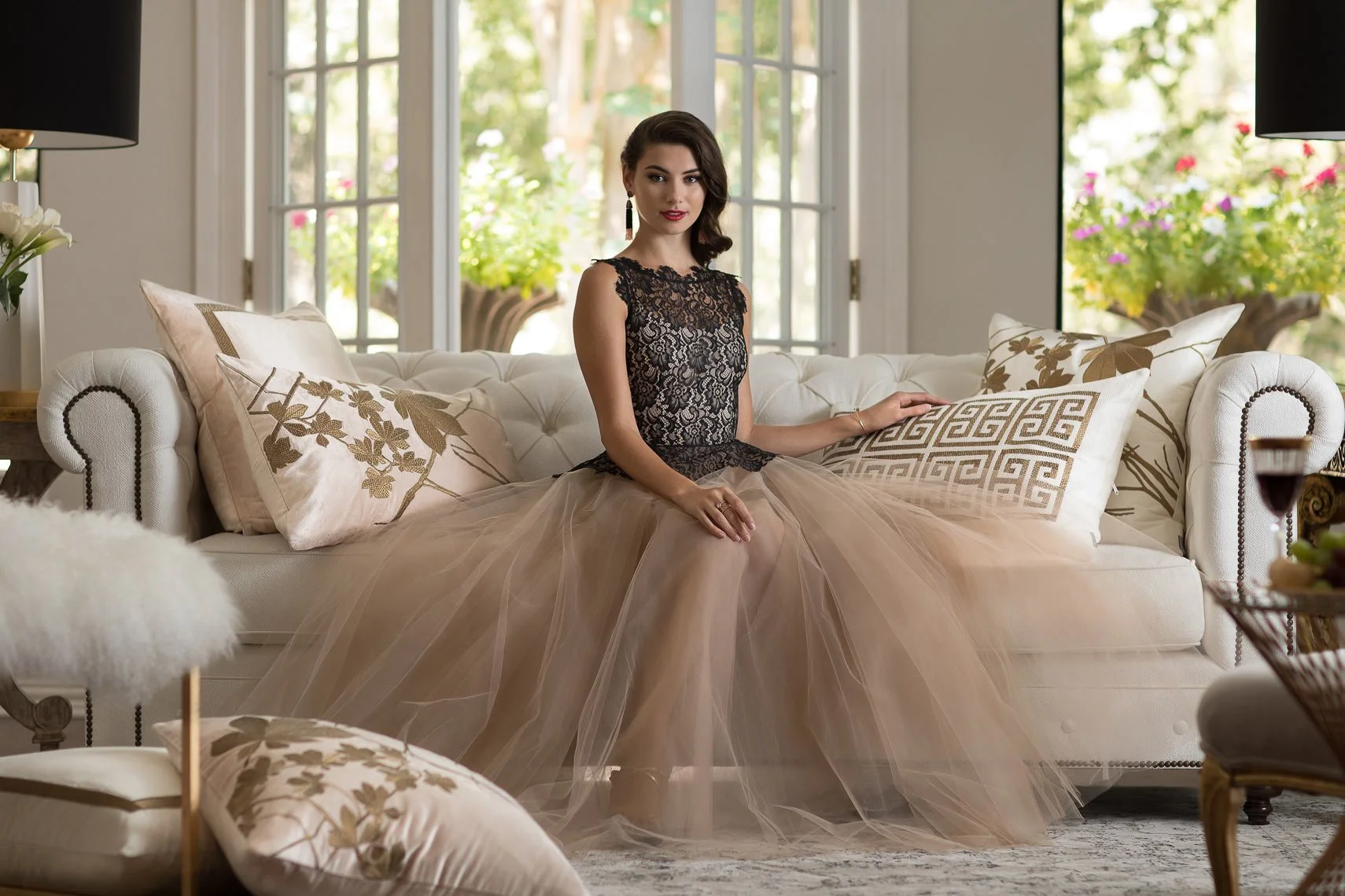 A woman in an elegant black lace top and beige tulle skirt sitting on a white tufted sofa with decorative cushions in a bright living room with large windows and floral outdoor view.