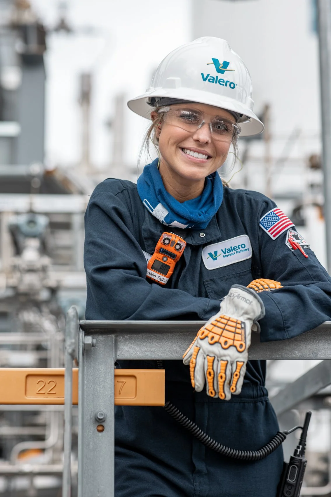 A smiling female worker wearing a white Valero helmet, safety glasses, and a dark uniform with patches, including an American flag and Valero logo, leaning on a metal railing inside a refinery or industrial facility.