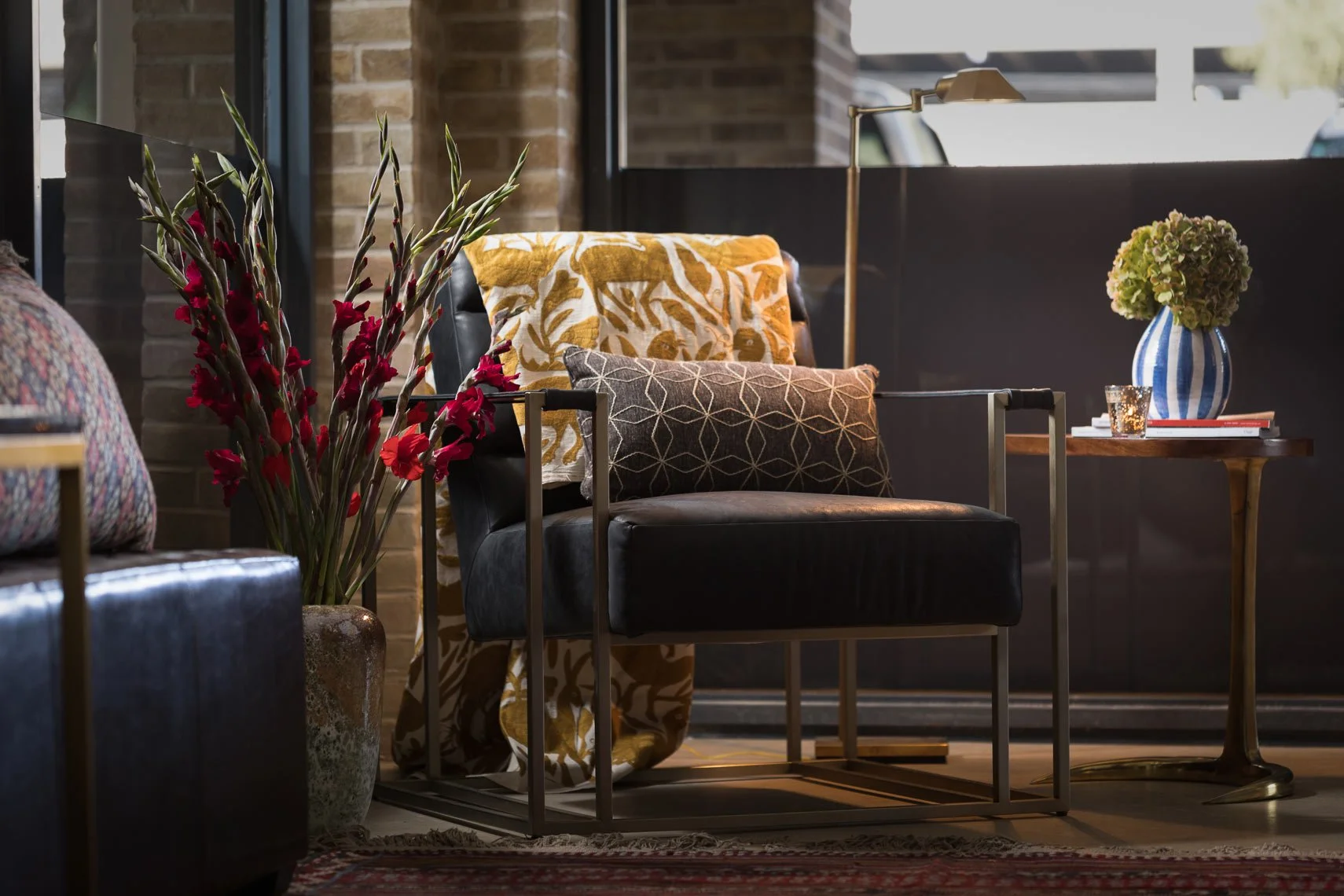 A cozy interior corner featuring a black leather armchair with patterned yellow and white cushions, a side table with a blue and white striped vase holding green hydrangeas, and a large vase with tall plants with red flowers, all near a brick wall an