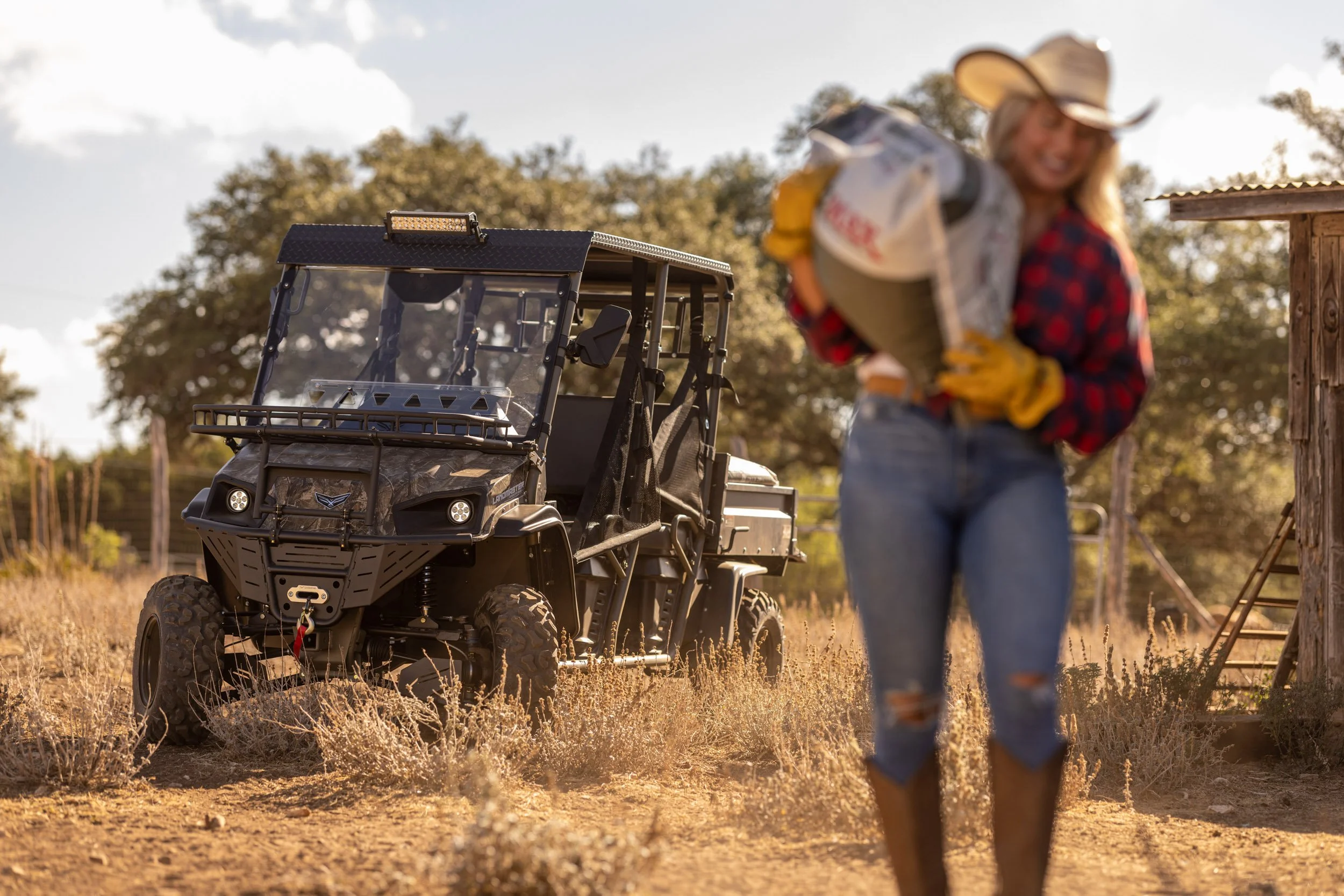 A woman wearing a cowboy hat, plaid shirt, and jeans standing outdoors in a rural setting, with a golf cart parked nearby on dry grass and trees in the background.