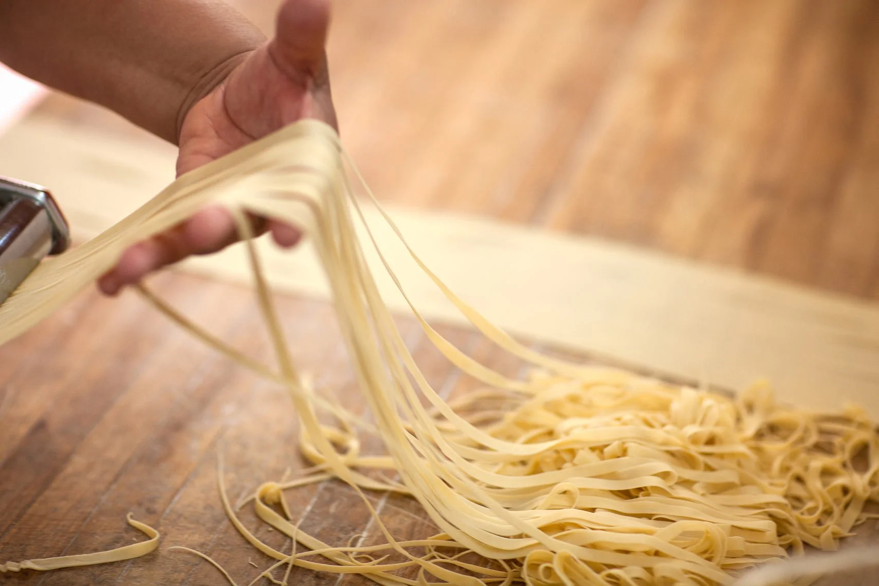 Close-up of a hand using a pasta machine to make fresh fettuccine, with pasta sheets and strands on a wooden surface.
