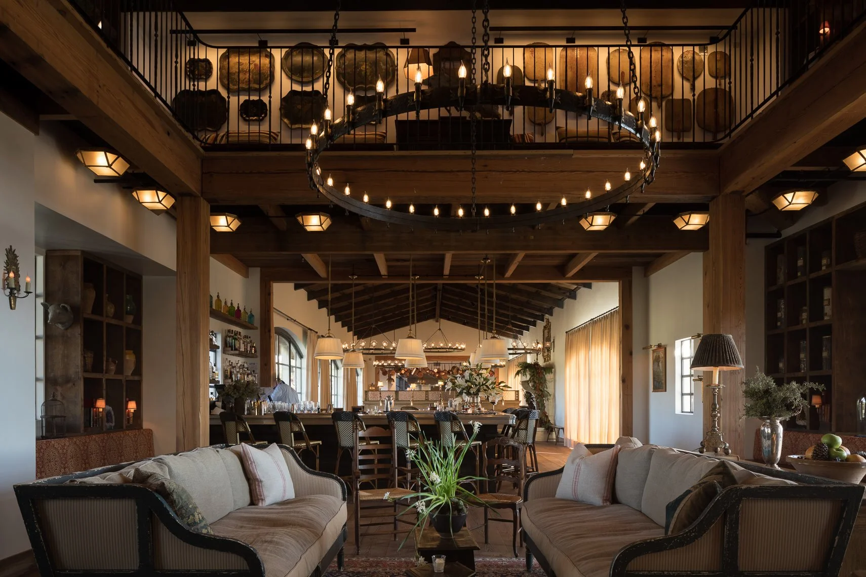 A spacious, cozy dining and living area with wooden beams, warm lighting, and modern rustic decor. The space features a large chandelier, tables, chairs, and decorative accents.