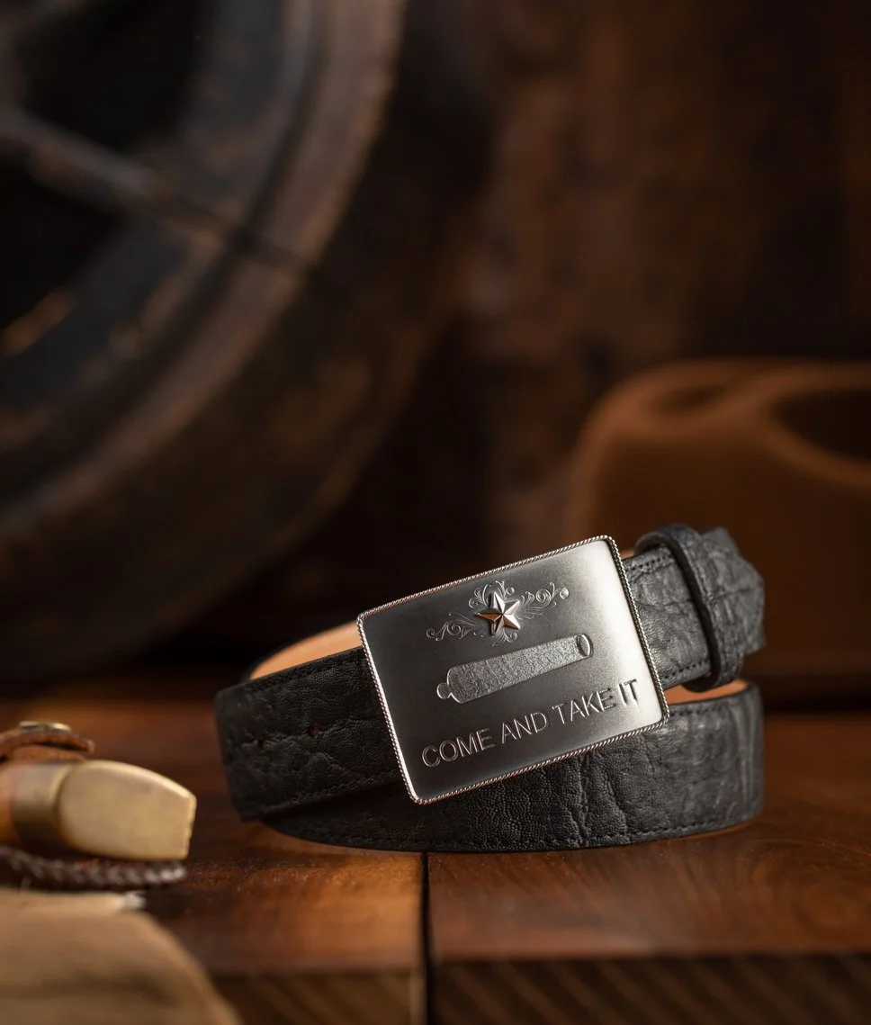 A black leather belt with a rectangular silver buckle engraved with a star, a scroll, and the text "COME AND TAKE IT" resting on a wooden surface.