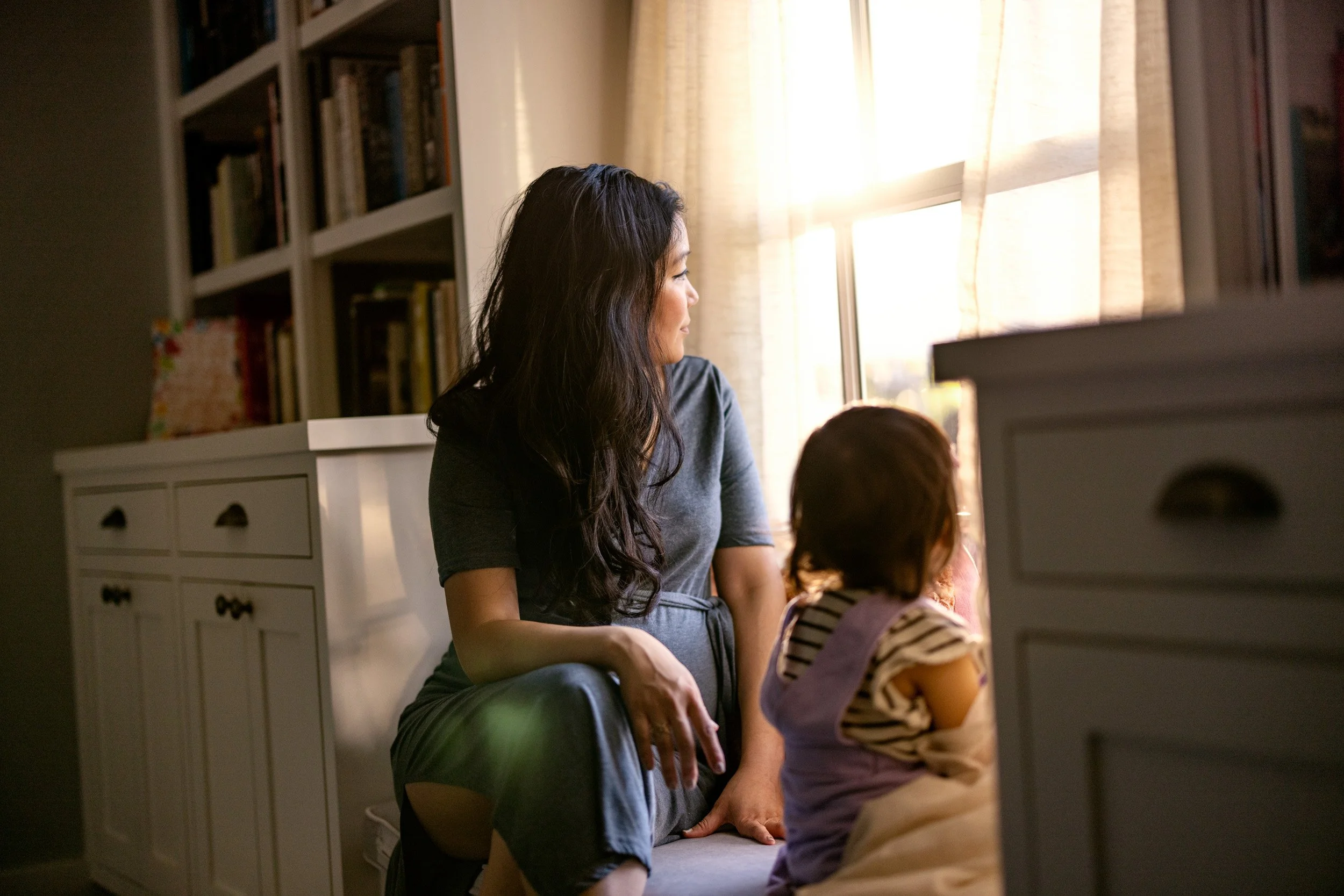 A woman and a young girl look out a sunny window in a cozy room with a bookshelf and white dresser.