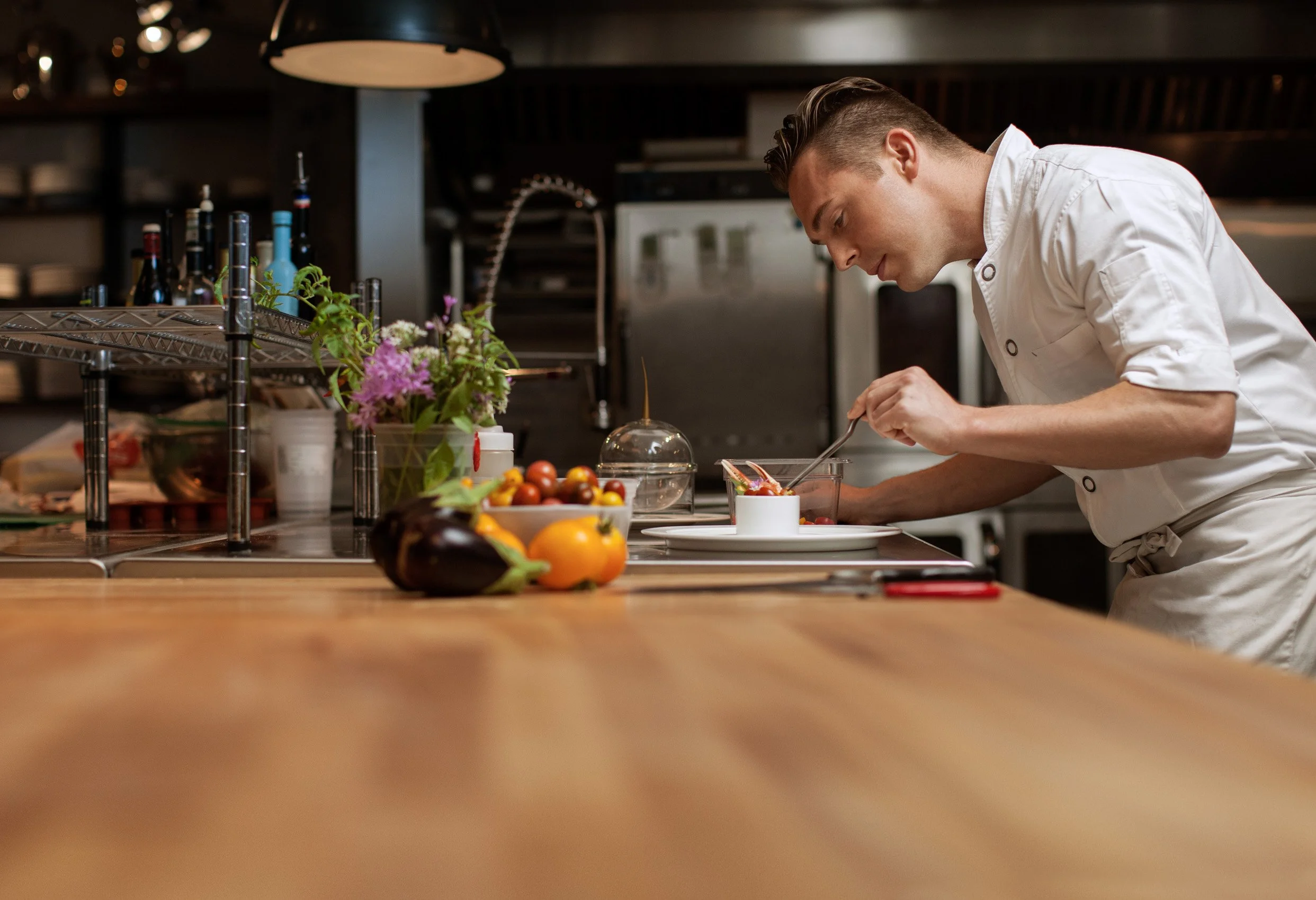 A chef in a white uniform is preparing food in a professional kitchen. The counter is filled with various fruits, vegetables, and flowers.