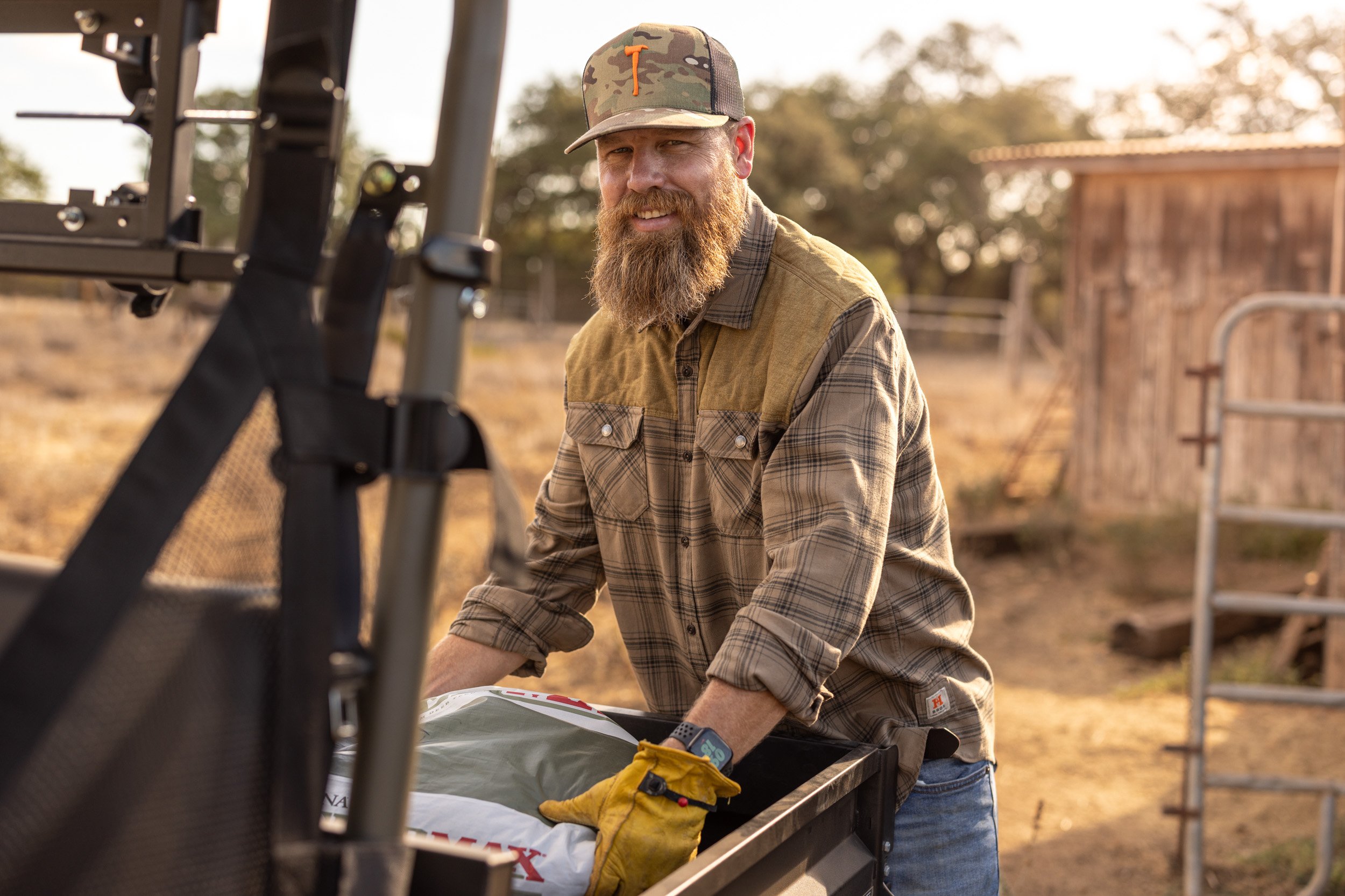 A man with a beard wearing a camo baseball cap and plaid shirt loading supplies onto a farm trailer outdoors during sunset.