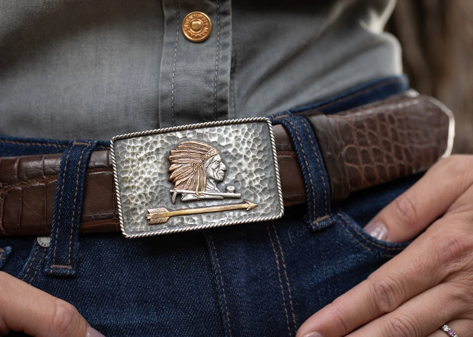 Close-up of a person wearing a brown leather belt with a silver and gold buckle featuring a Native American chief in profile and an arrow. The person is dressed in blue jeans and a gray shirt.