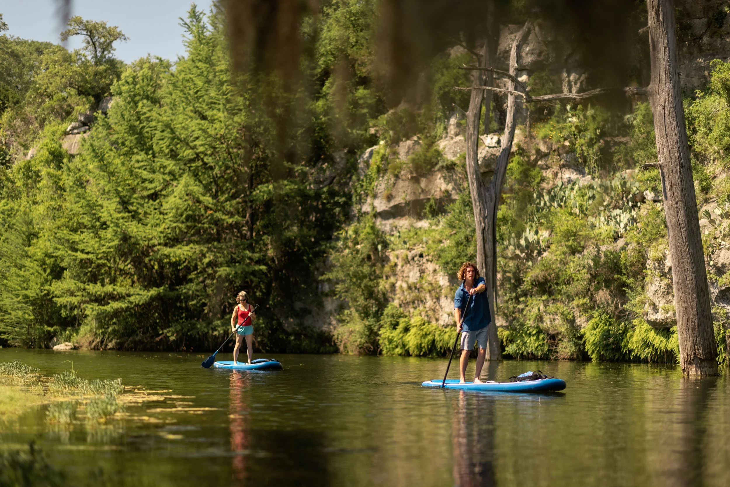 Two people paddleboarding on a calm river surrounded by lush green trees and rocky cliffs.