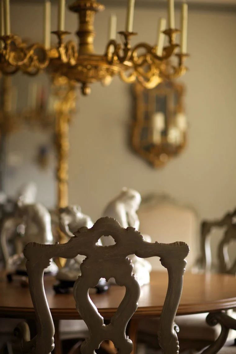 Close-up of an ornate antique dining chair back with a carved design in front of a wooden dining table, with decorative porcelain lions and a gold chandelier hanging above in a traditionally decorated room.