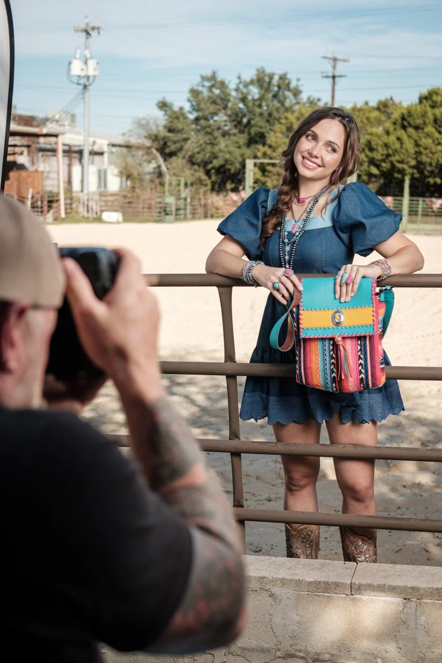 A young woman wearing a blue dress with puffed sleeves and cowgirl boots posing for a photo behind a metal fence at an outdoor setting. She has long hair styled in a braid, is smiling, and looks at the photographer. She is accessorized with colorful 