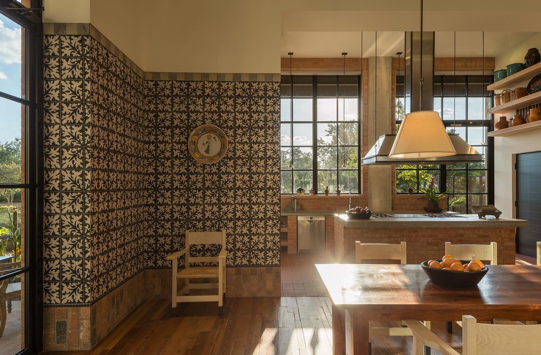 Sunlit kitchen with patterned tile wall, wooden dining table with a bowl of oranges, large window with plants on the sill, and open shelving with pottery