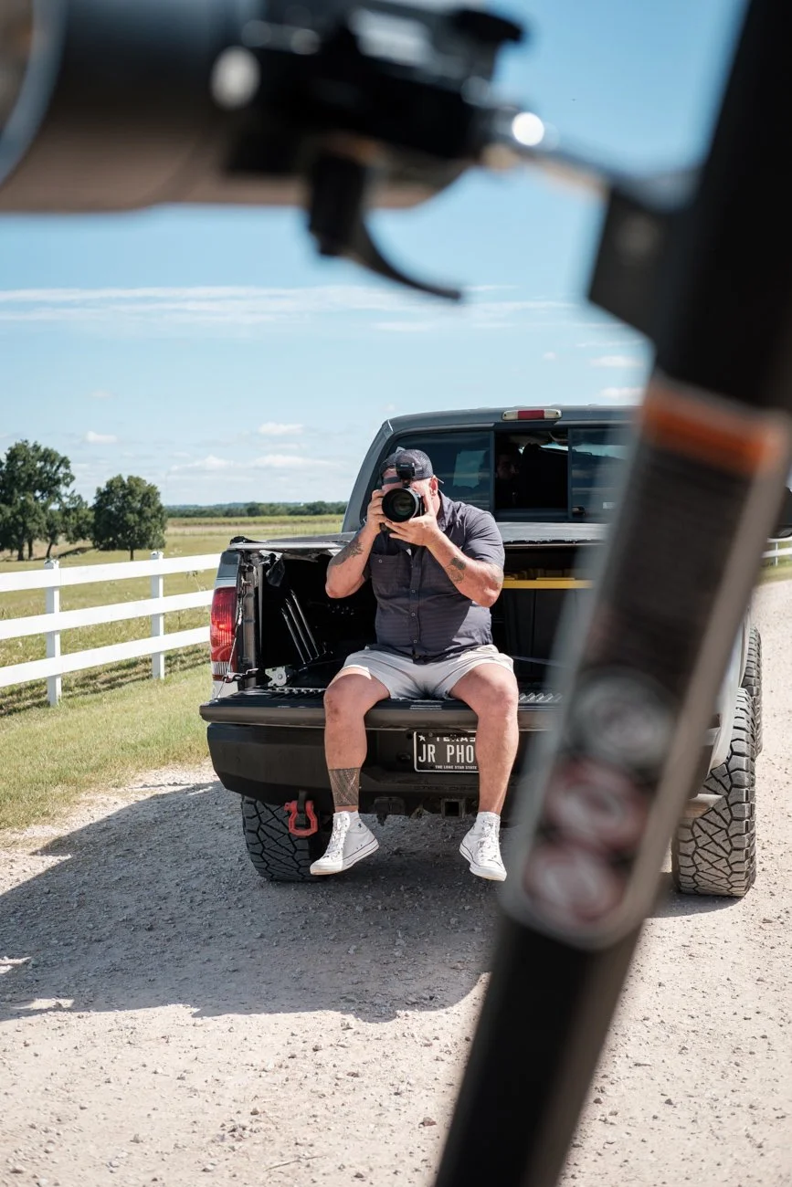 A man sitting on the tailgate of a black pickup truck, taking a photo with a camera. The photo is reflected in a mirror or another reflective surface in the foreground. The man is wearing a dark shirt, shorts, white sneakers, and a cap. The backgroun