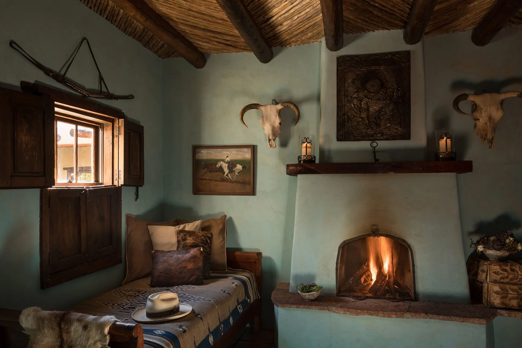 Cozy room with a lit fireplace, wooden ceiling beams, two skull mounted on the wall, a window with wooden shutters, a framed painting of a horse, and a bed with pillows, a hat, and a fur blanket.