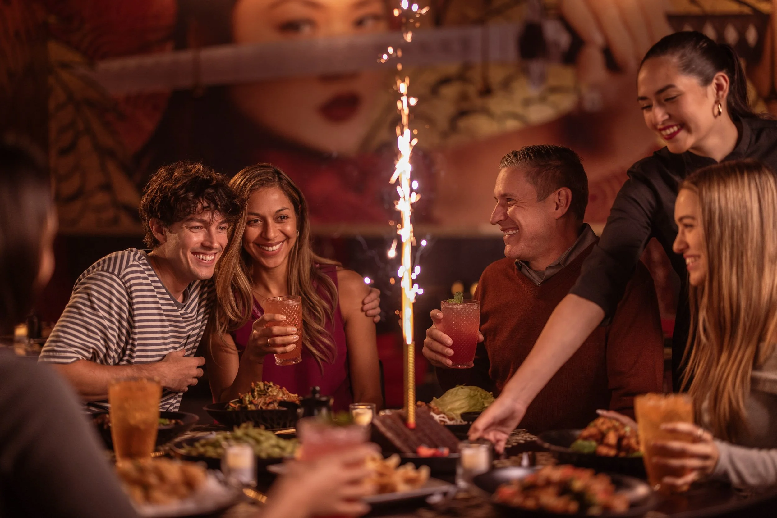 People celebrating a birthday with a cake, sparklers, and drinks in a cozy restaurant or bar setting, smiling and enjoying each other's company.