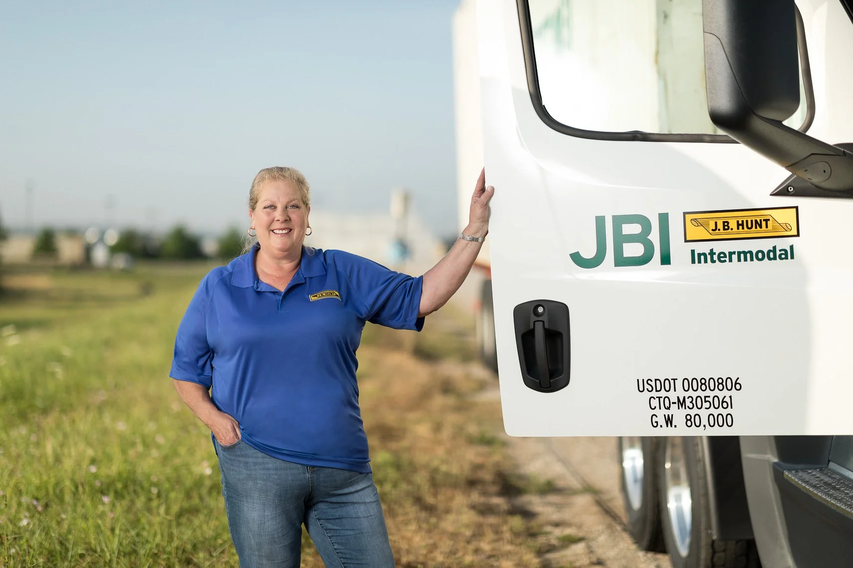A woman in a blue polo shirt standing outdoors next to a large white vehicle with company branding, smiling and resting her hand on the vehicle.