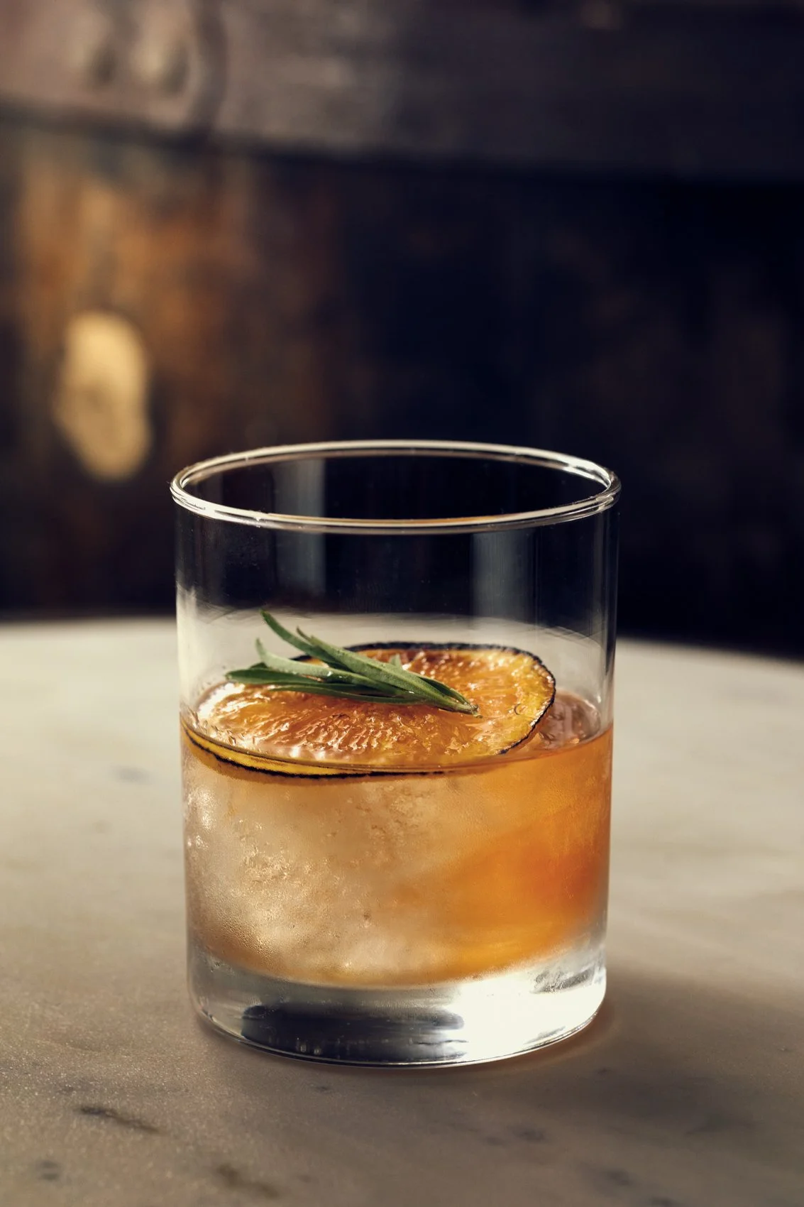 A clear glass with a cocktail garnished with a slice of orange and a sprig of rosemary on top, sitting on a light-colored surface.