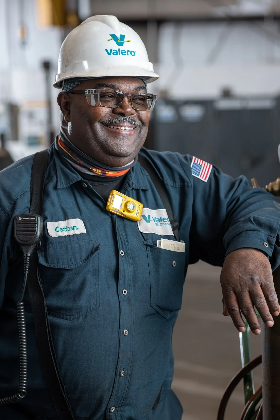 A smiling male utility worker wearing safety glasses, a white hard hat with the Valero logo, a navy blue uniform with name tag 'Cotton', and a yellow safety device around his neck. He is standing in an industrial setting with equipment and wiring vis