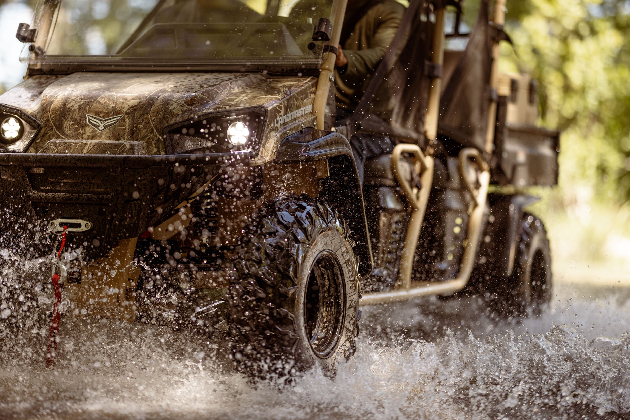 A camouflage all-terrain utility vehicle driving through muddy water, splashing water around with large black tires and a front light on, with a person in a green jacket steering. Trees and sunshine in the background.