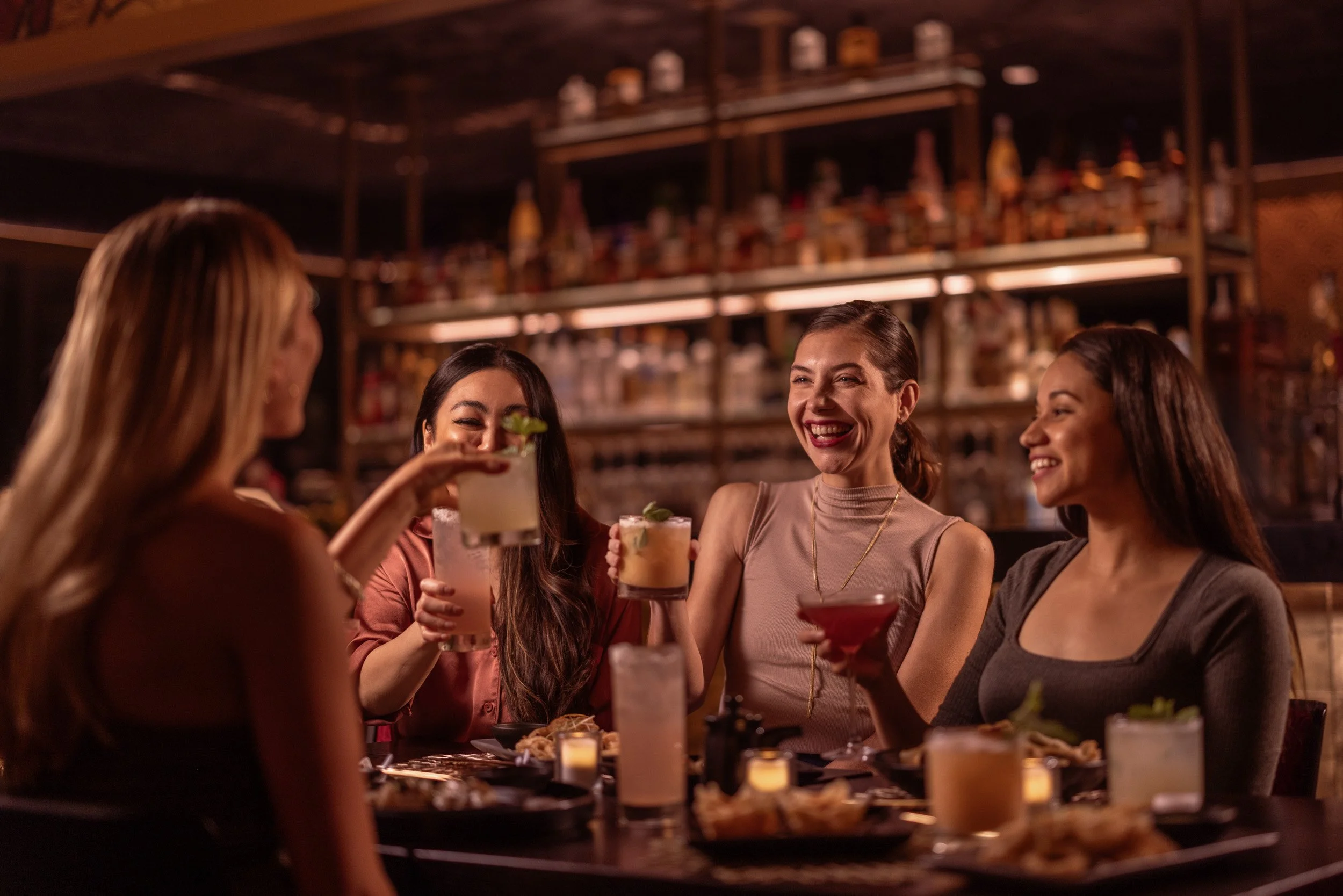 A group of five women enjoying drinks and conversation at a restaurant or bar, with shelves of bottles and glasses in the background, and candles on the table.