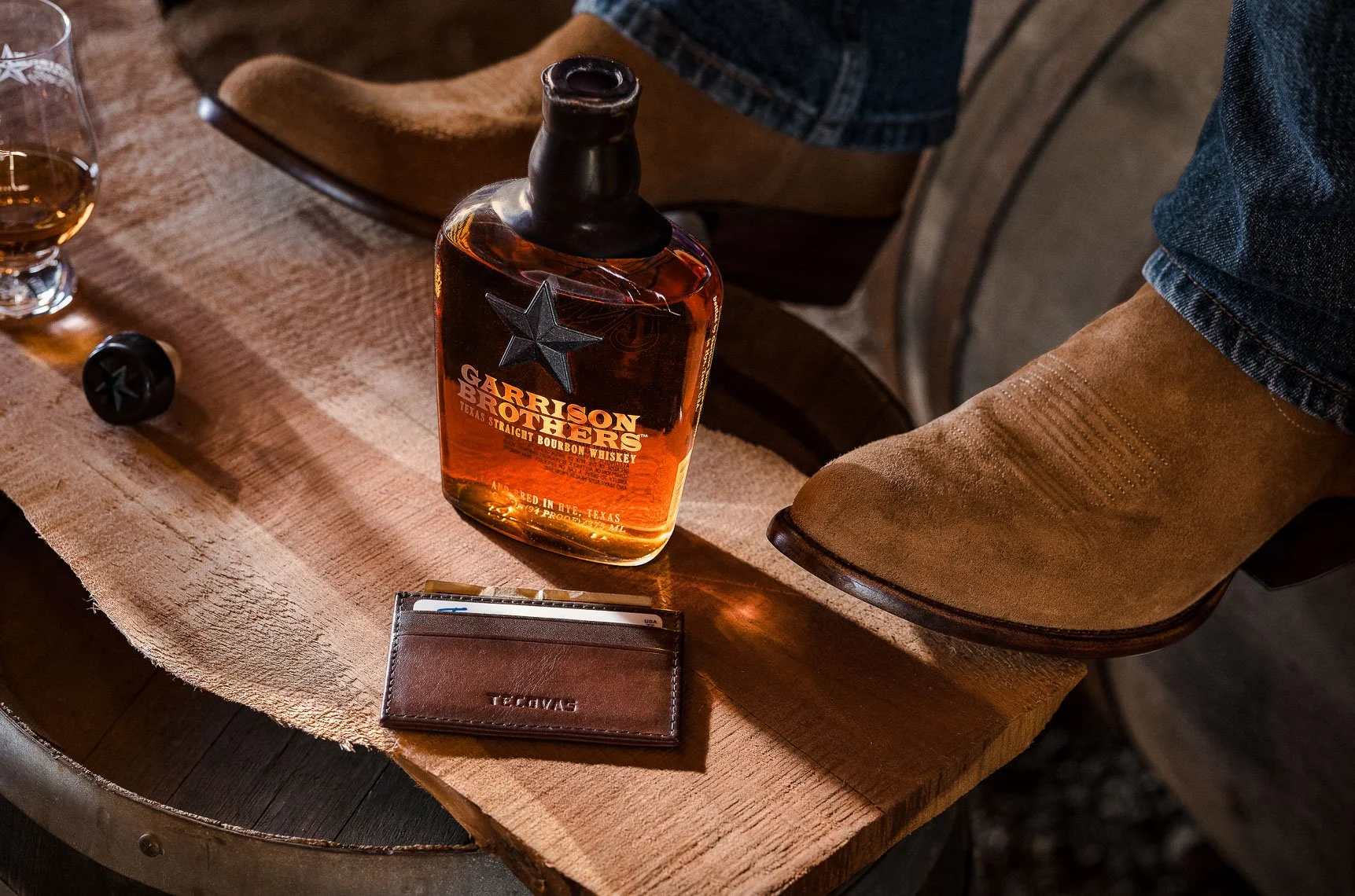 A bottle of Garison Brothers Texas Straight Bourbon Whiskey on a wooden table next to a glass of whiskey, a wallet, and a cowboy boot.