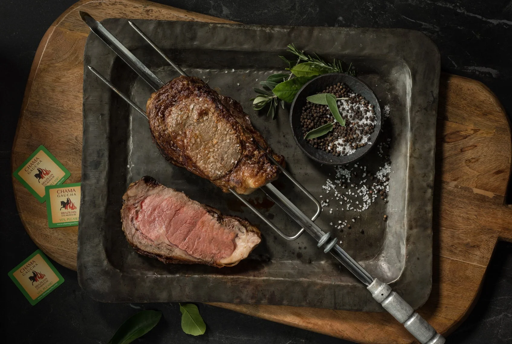 A tray with two cooked cuts of steak on a metal rack, garnished with herbs and sea salt, with a syringe, salt, and pepper nearby, on a wooden board.