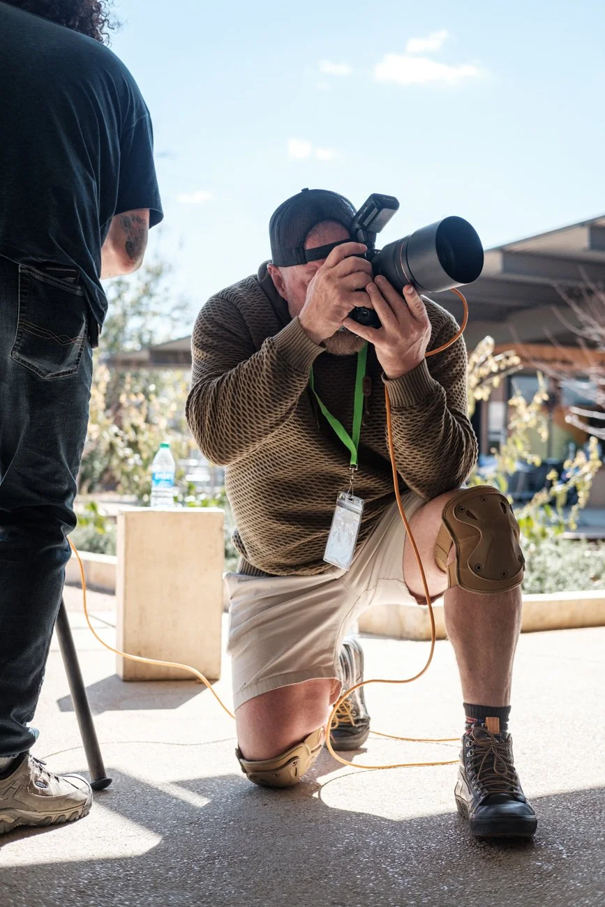 A man crouching with one knee on the ground, holding a camera with a large telephoto lens, taking a photograph outdoors under a clear blue sky. He wears a brown sweater, beige shorts, a black cap, and protective knee gear. Another person stands nearb