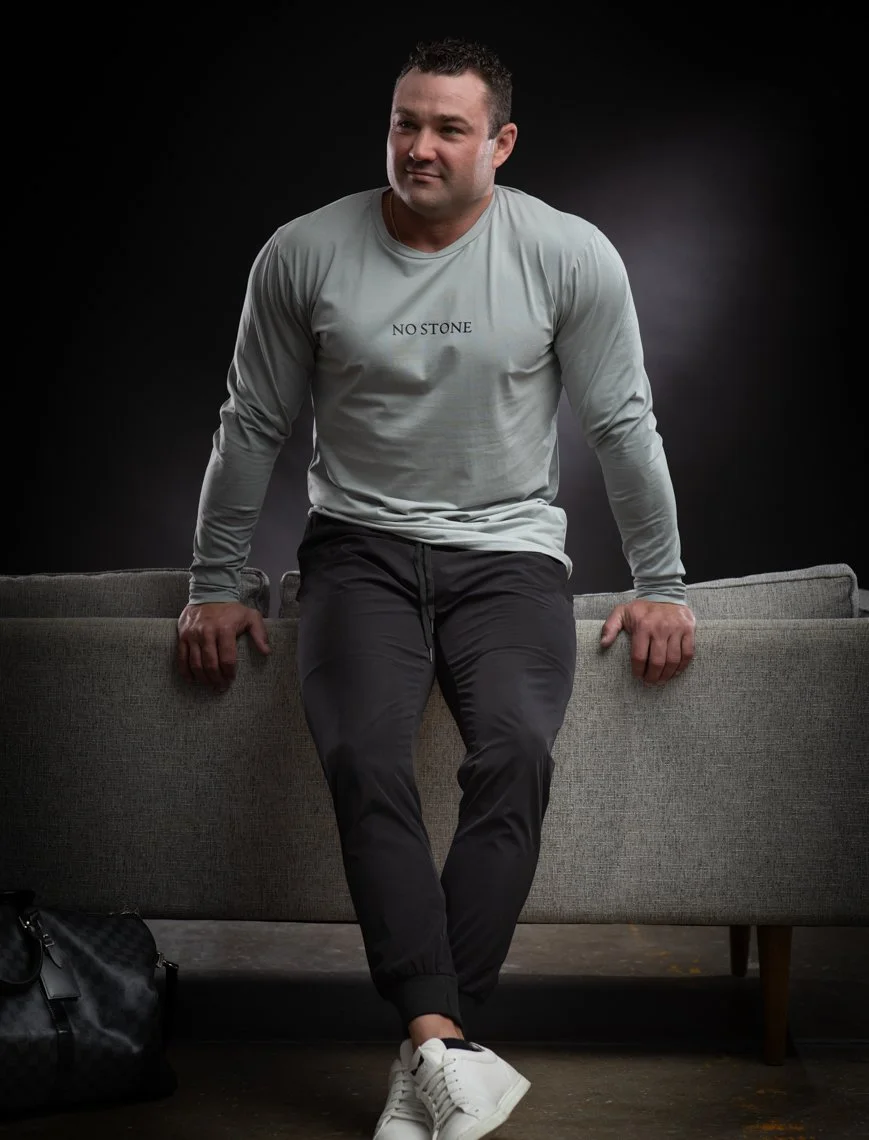 A man is sitting on a gray sofa in front of a black background, wearing a light gray long-sleeve shirt with the words 'NO STONE' printed on it, black pants, and white sneakers.