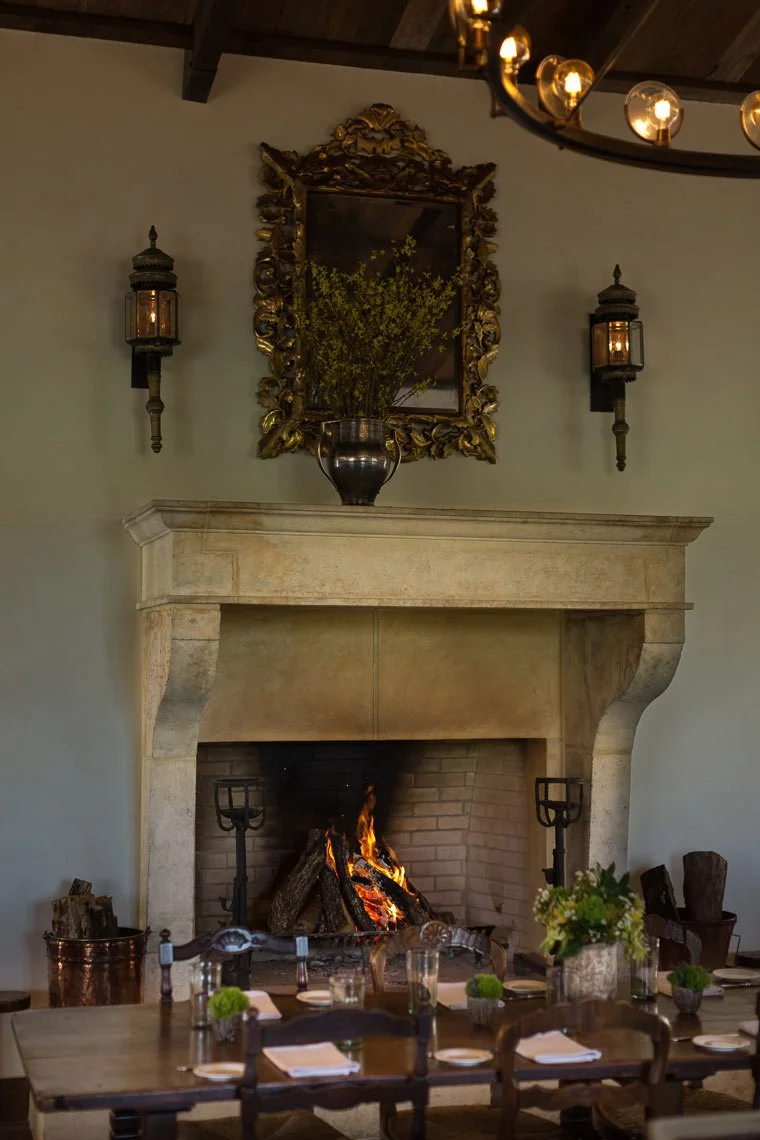 Interior of a cozy dining room with a lit fireplace, a large mirror above it, and vintage wall lamps. A table set with napkins, plates, and glasses. Decor includes a vase with flowers and firewood.