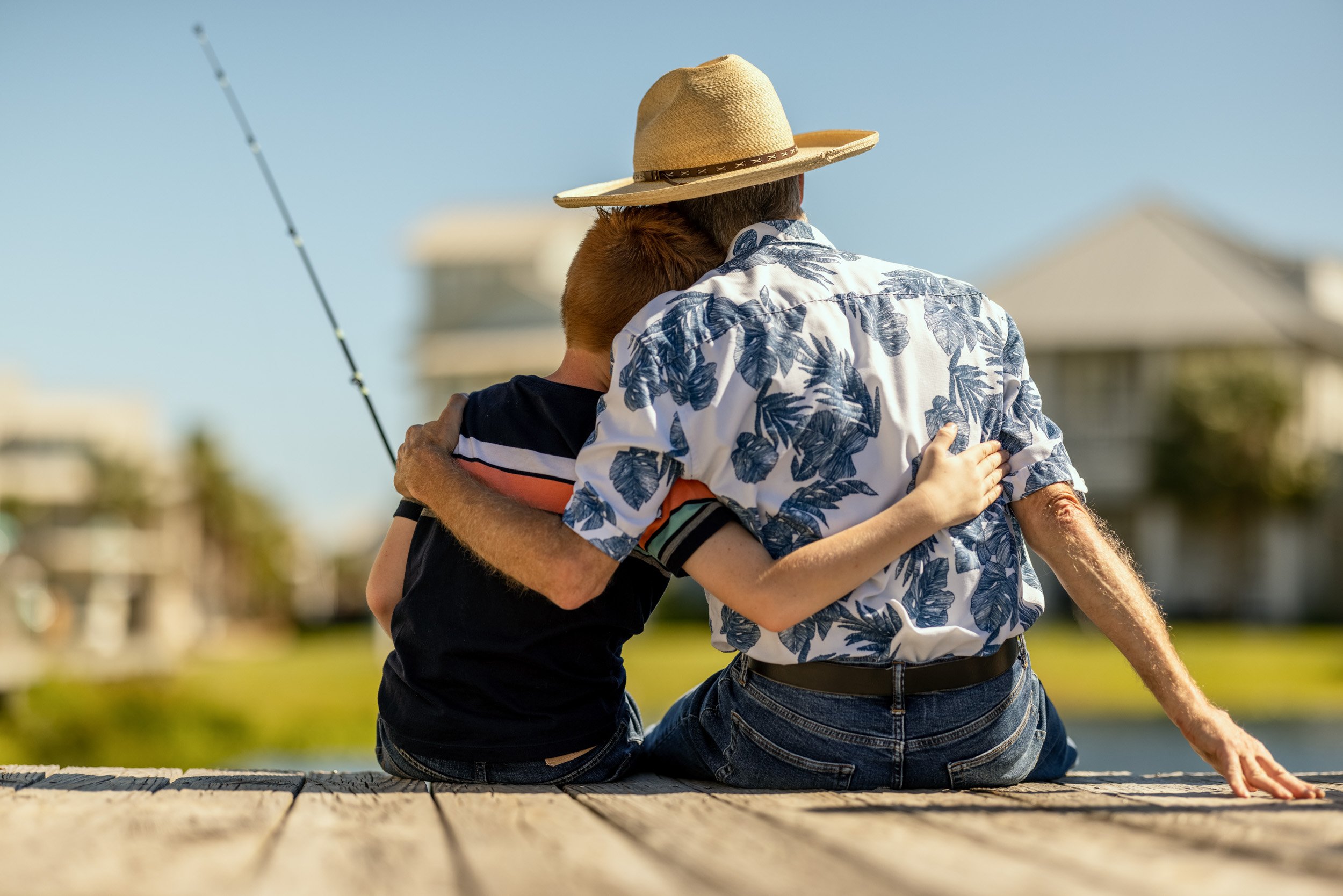 Two people sitting on a dock hugging, one wearing a straw hat and a floral shirt, the other in dark clothing, with a fishing pole nearby and houses in the background under a clear sky.