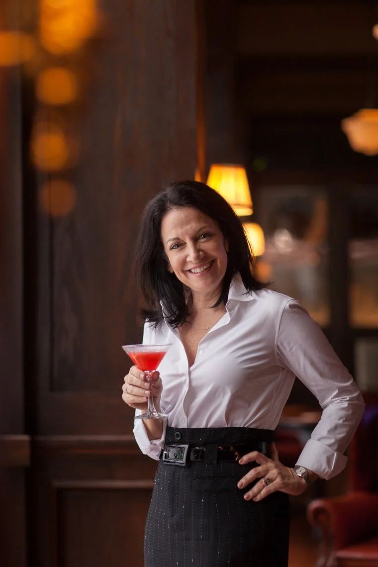 A woman smiling at the camera while holding a pink cocktail in a restaurant or bar with warm lighting and wooden decor.