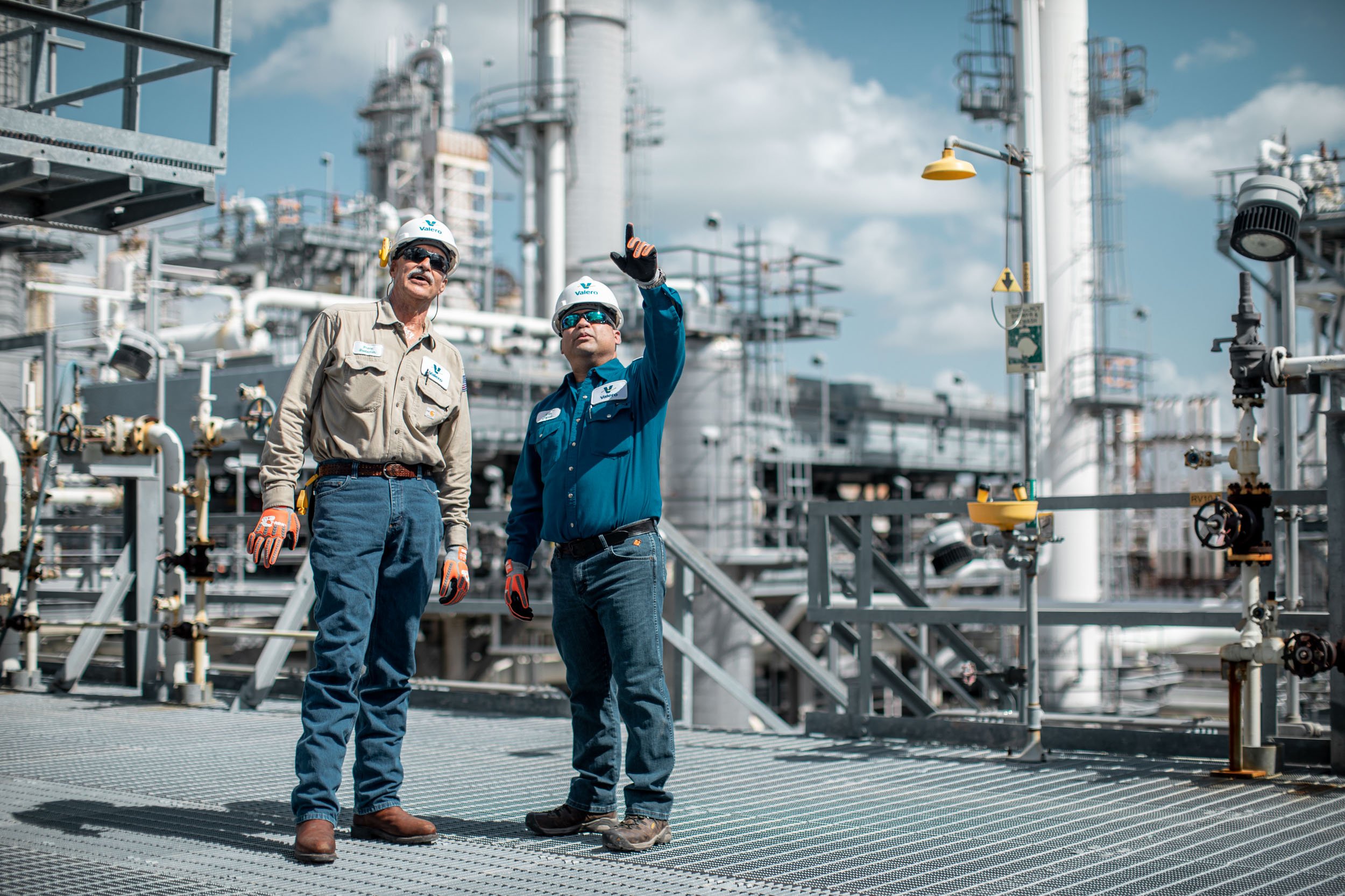 Two male industrial workers wearing hard hats and safety gear, inspecting equipment at an oil refinery or chemical plant with pipes and structures in the background.