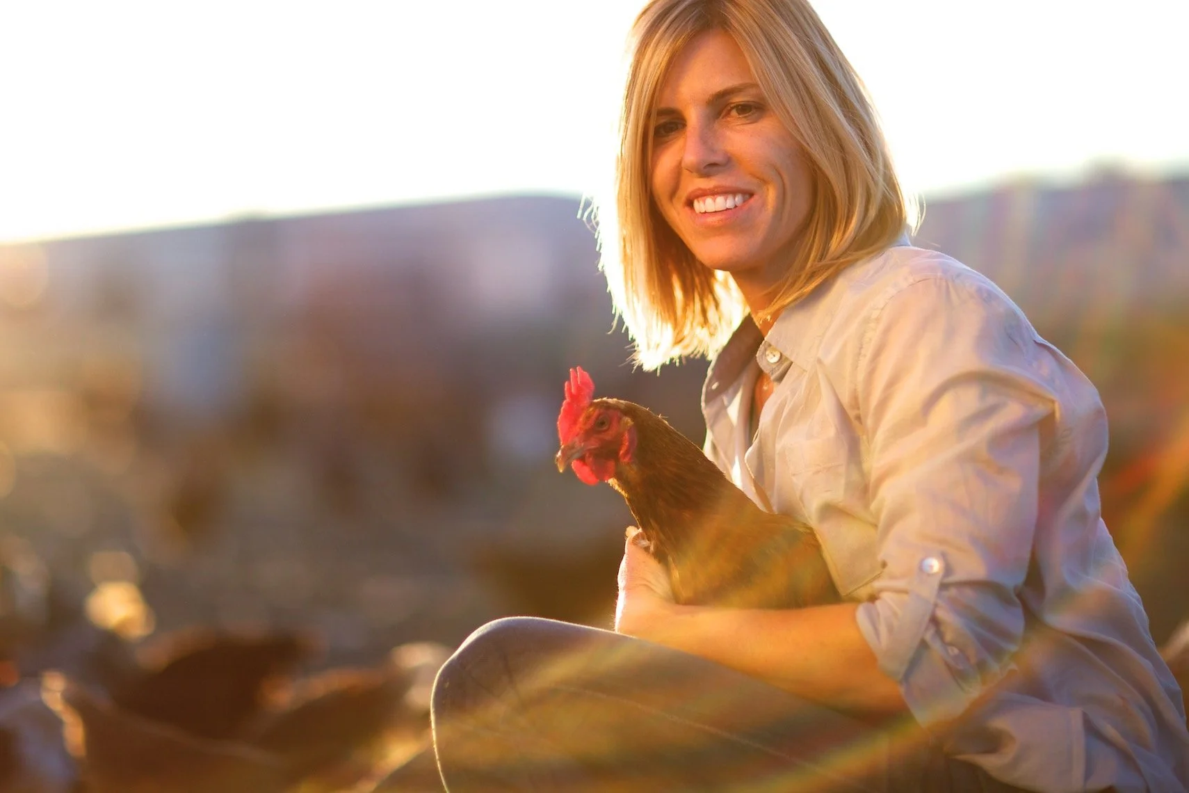 A woman with shoulder-length blonde hair holding a chicken and smiling outdoors during sunset.