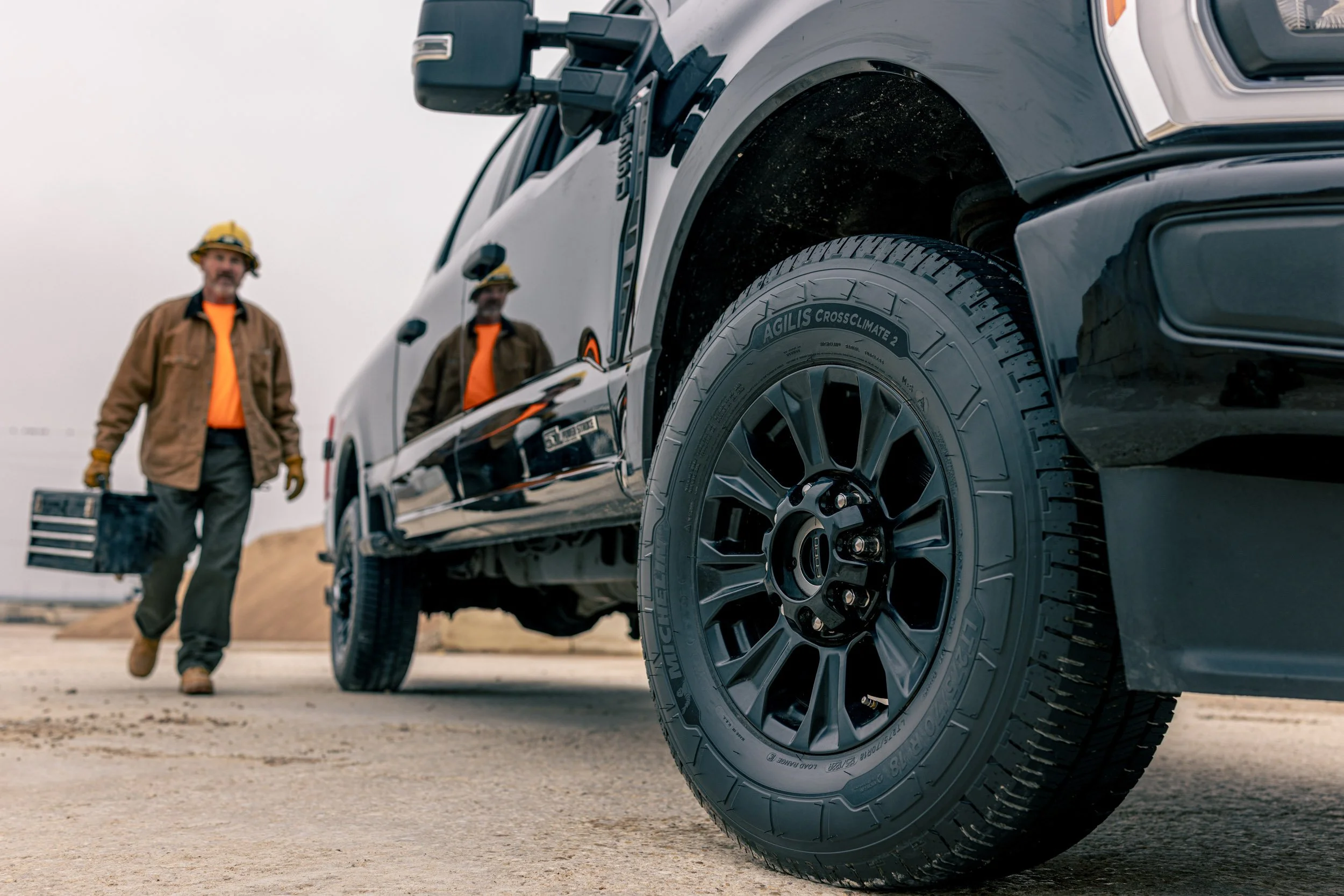 Close-up of a tire on a vehicle with two workers in hard hats and safety jackets walking in the background on a construction site or off-road area.