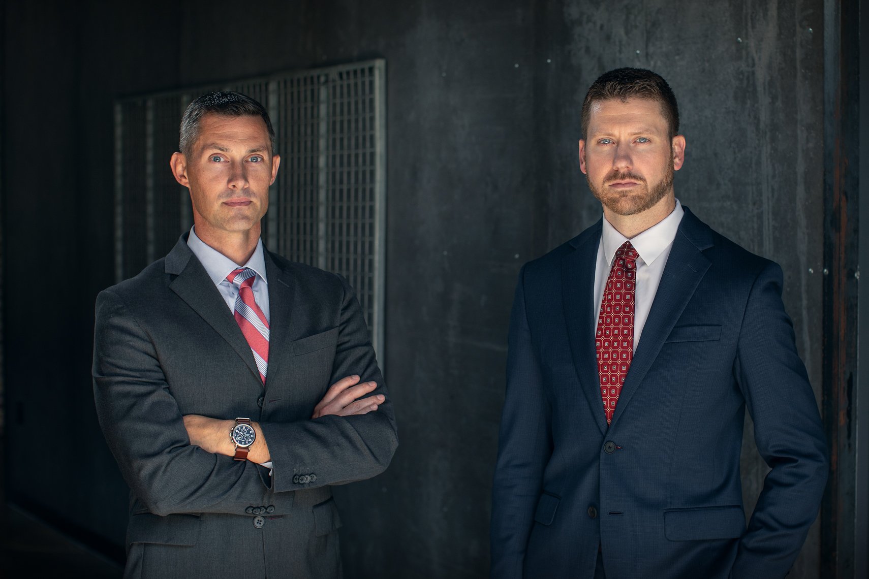 Two men in business suits standing against a dark wall, with serious expressions and arms crossed.
