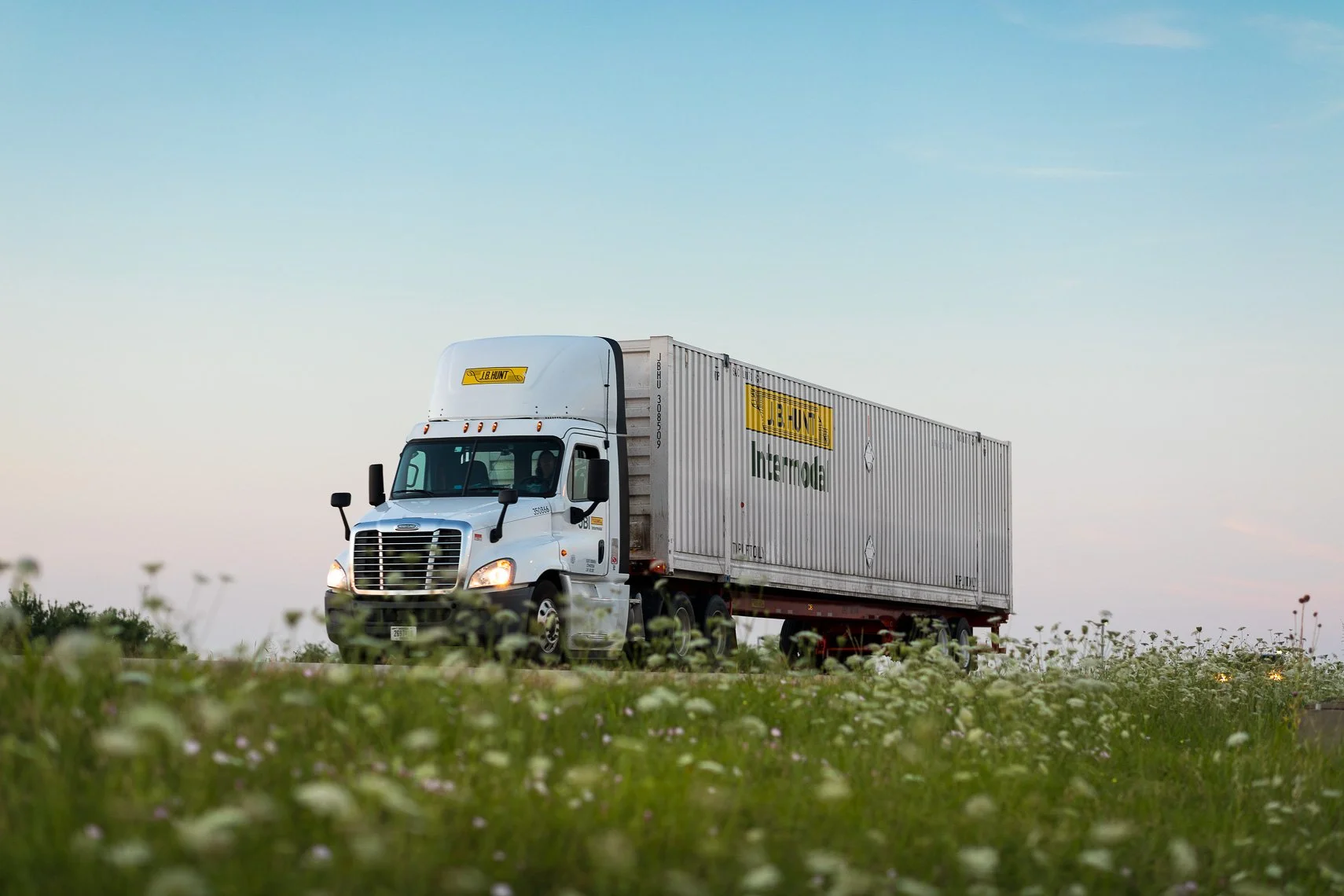 A white semi-truck with a large trailer traveling through a grassy field at sunset with a clear sky.
